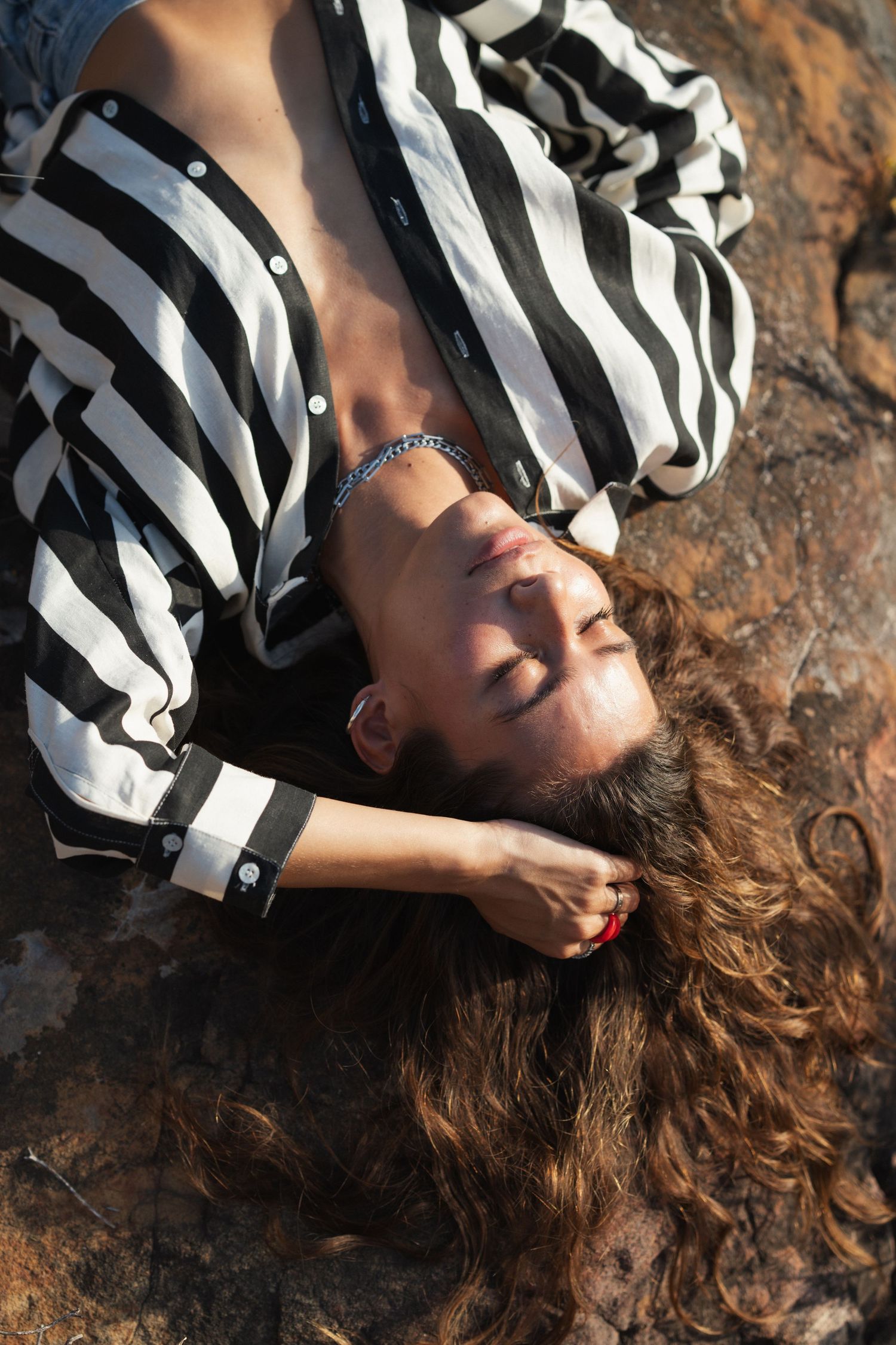Woman with curly hair lying on rocks, eyes closed, wearing a black and white striped shirt with a bracelet.