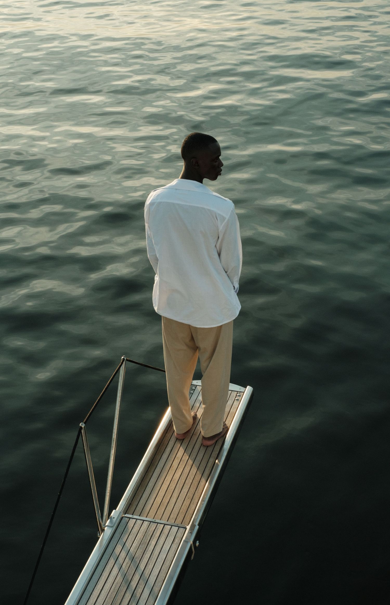 A man in a white shirt and beige pants stands on a wooden dock, gazing out over calm, dark water.
