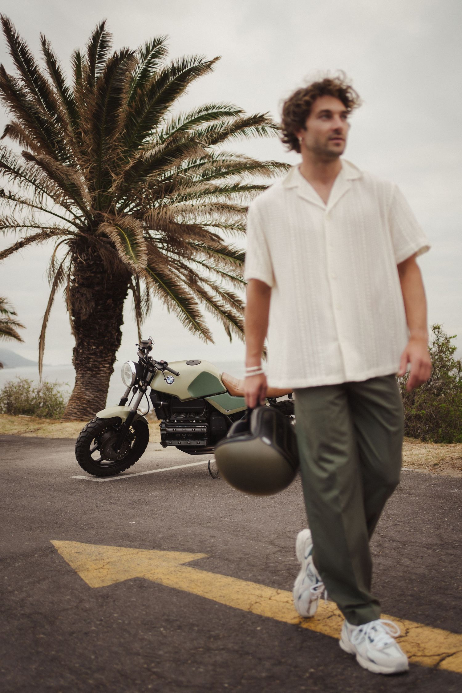 Man in white shirt and green pants holds helmet near motorcycle and palm tree on overcast day.