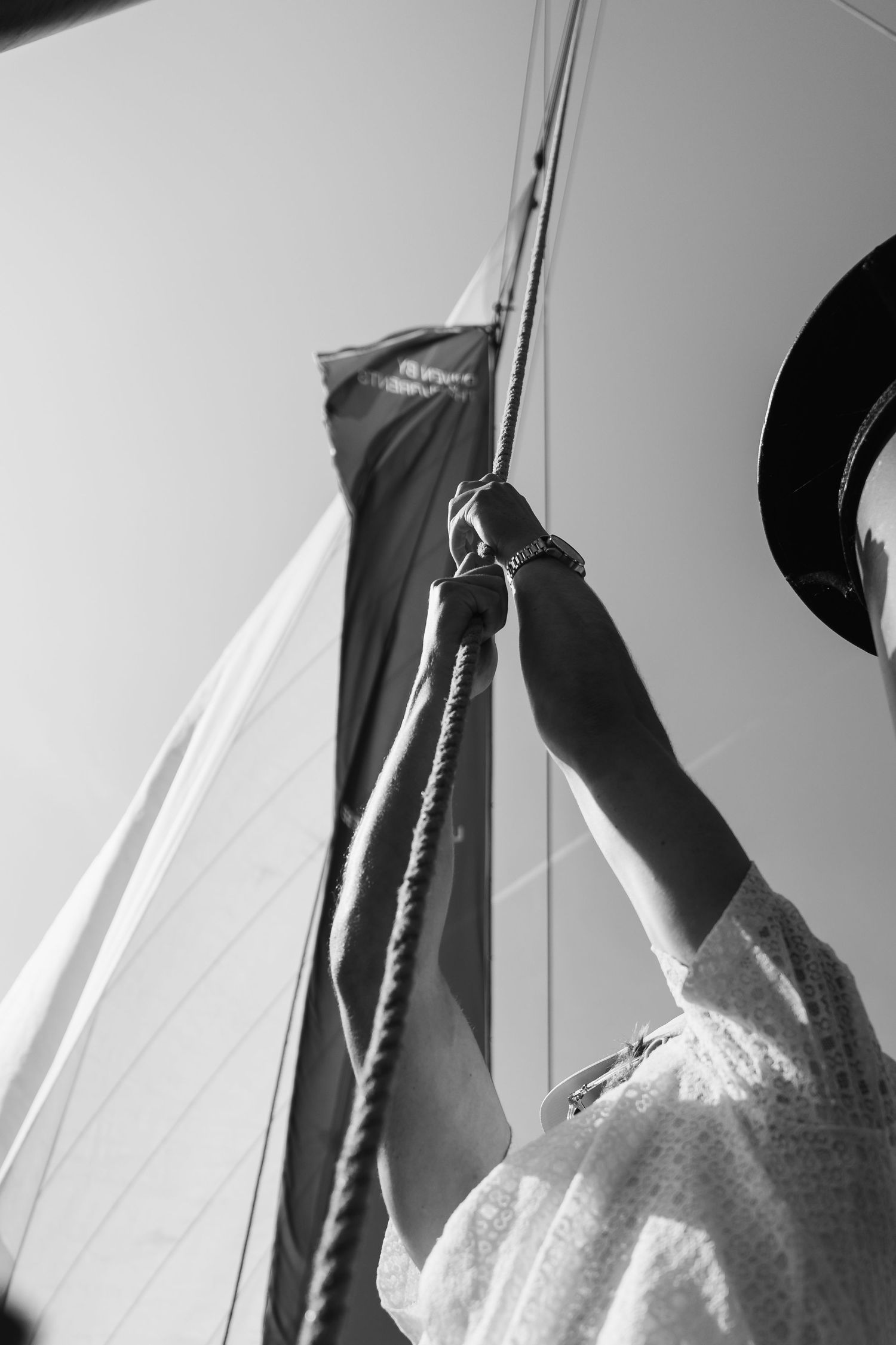 Black and white photo of a woman in a hat holding sailboat rigging ropes against a clear sky.