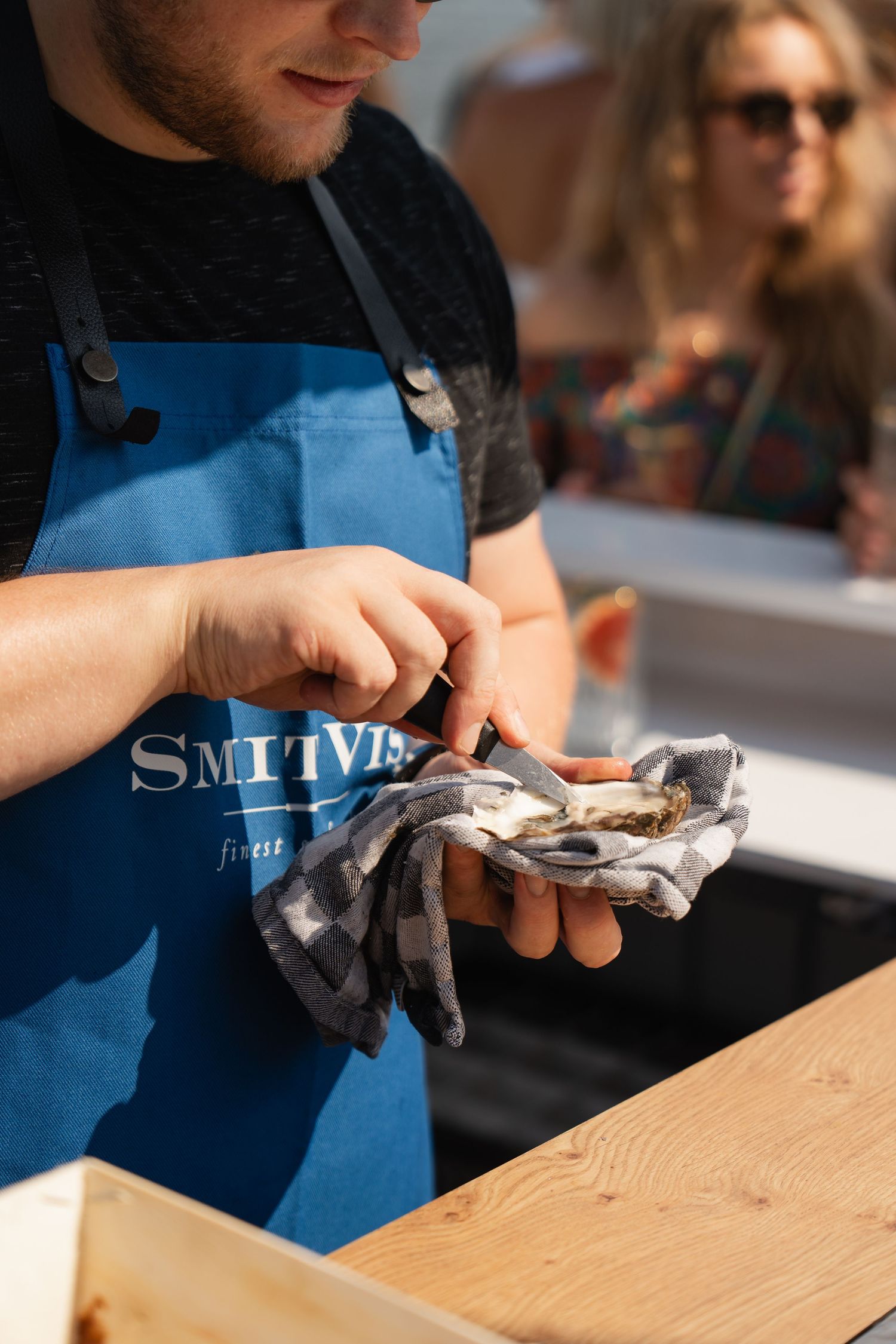 A man in a blue Smitty's apron shucking a fresh oyster with a cloth and knife outdoors.