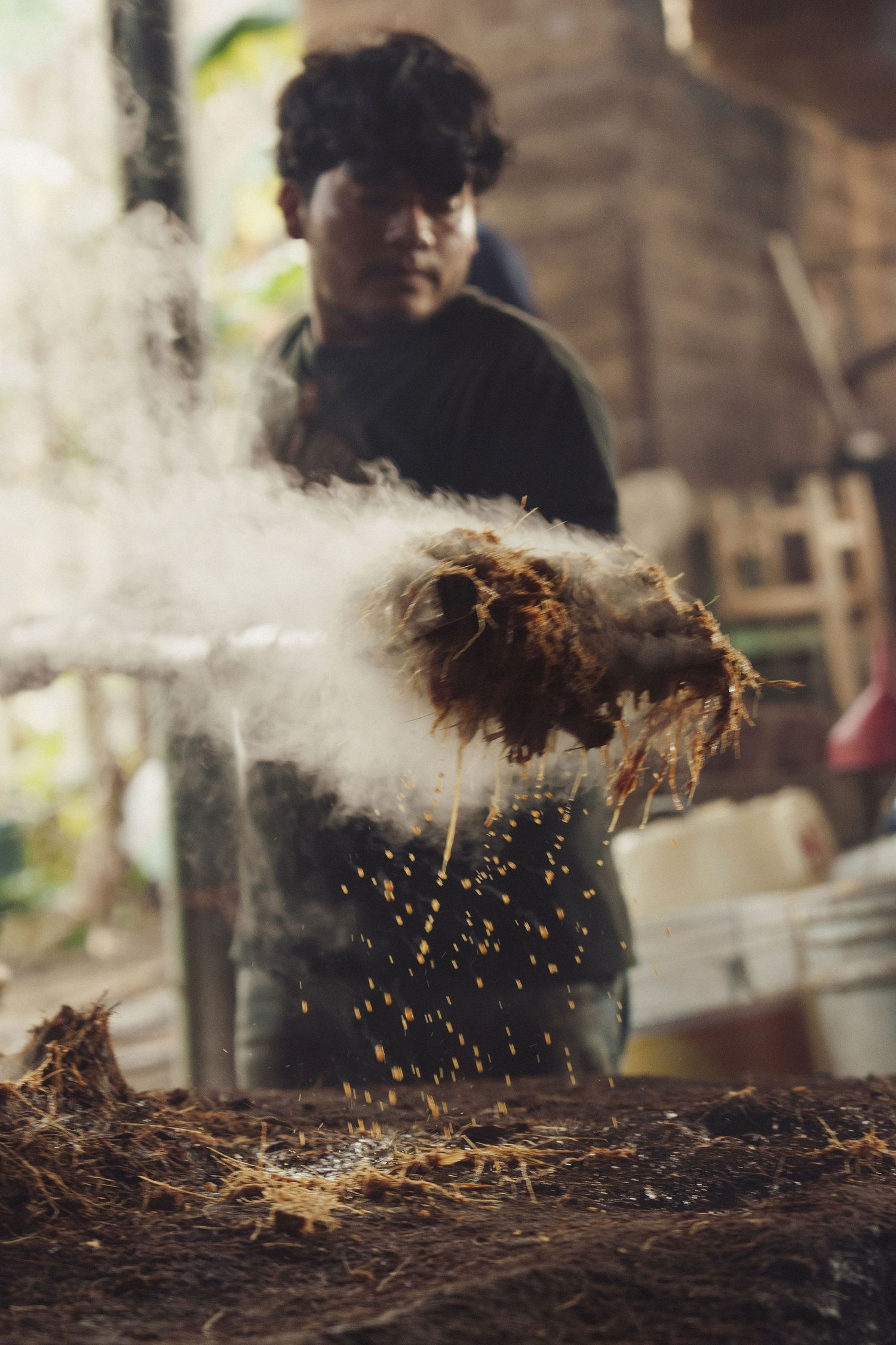 A man processes coconut fiber, creating dust clouds in a rustic workshop setting.