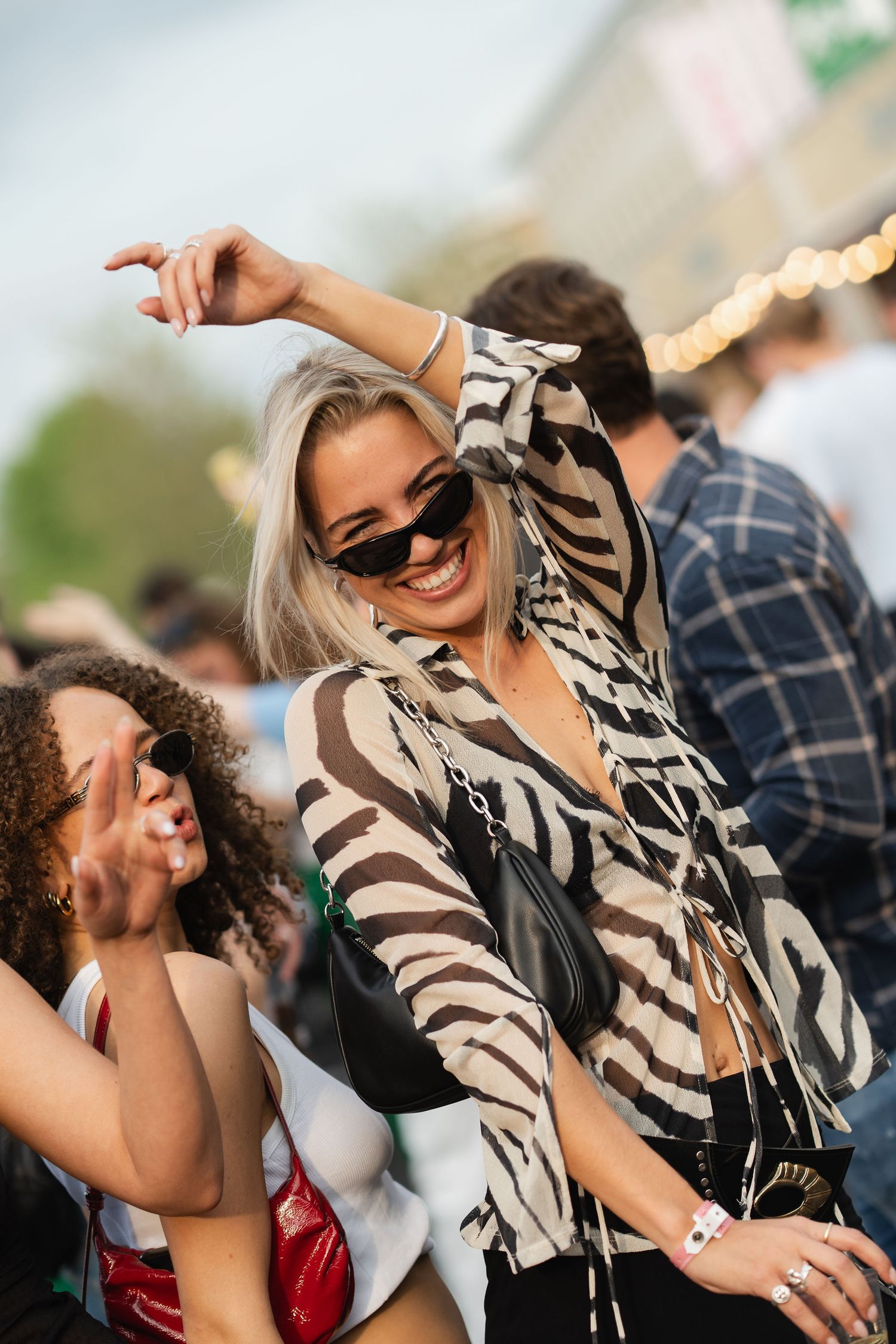 A smiling blonde woman in a zebra-print top and sunglasses dances joyfully at an outdoor festival.