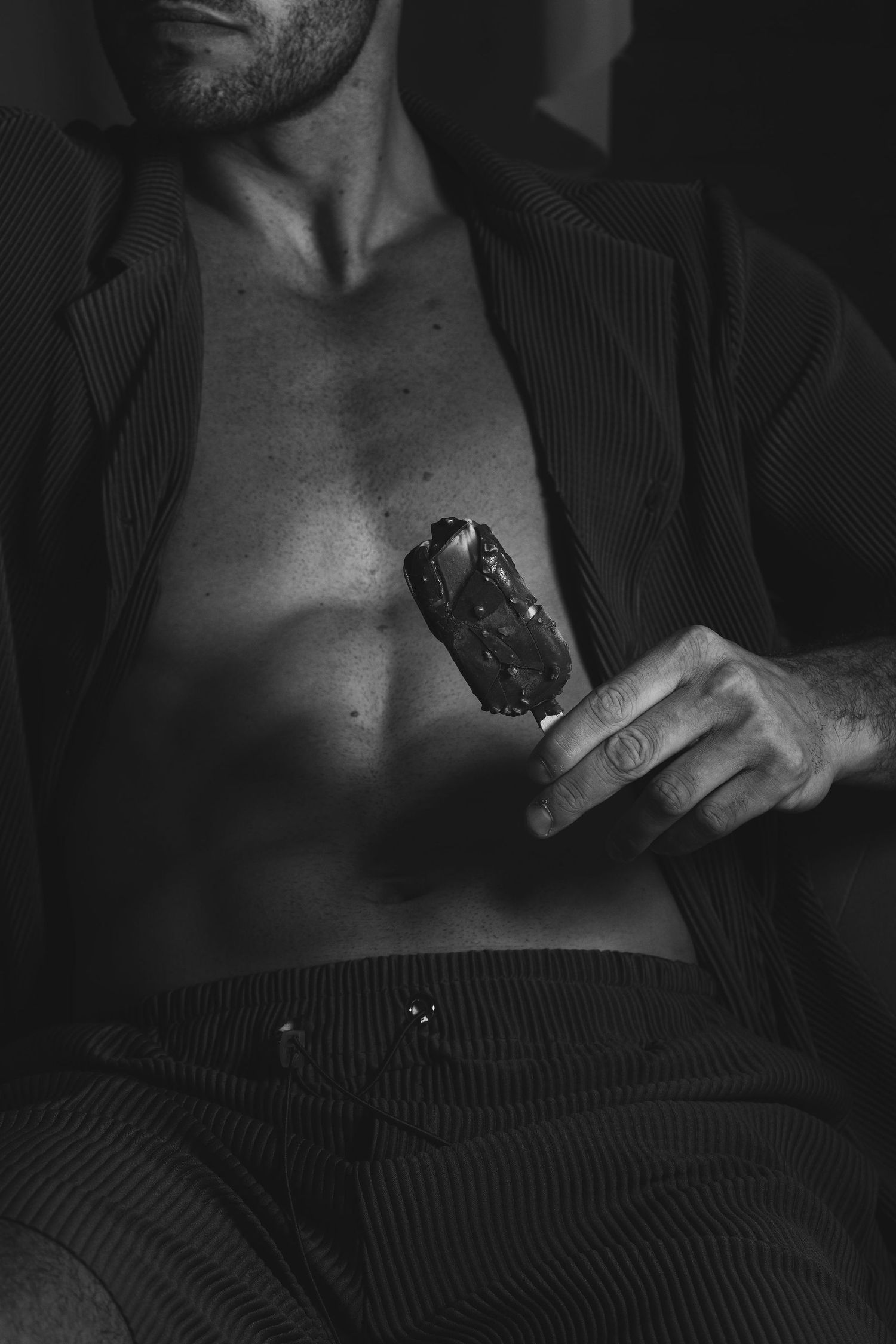 Black and white photo of a shirtless muscular man in an open suit jacket holding a chocolate ice cream bar.