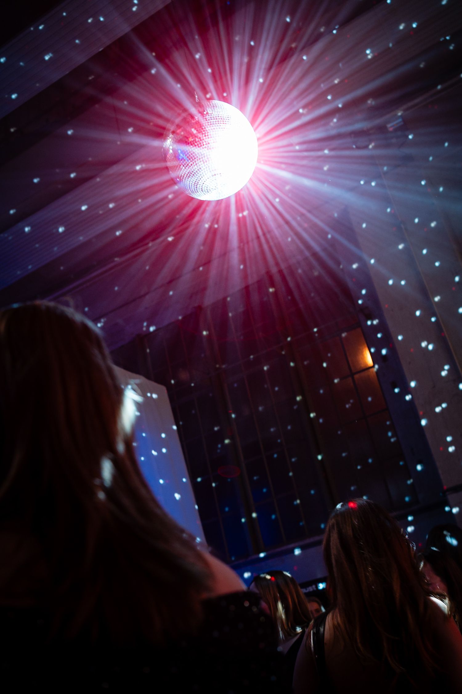 A glowing disco ball casting colorful light rays across a darkened dance floor with silhouetted partygoers below.