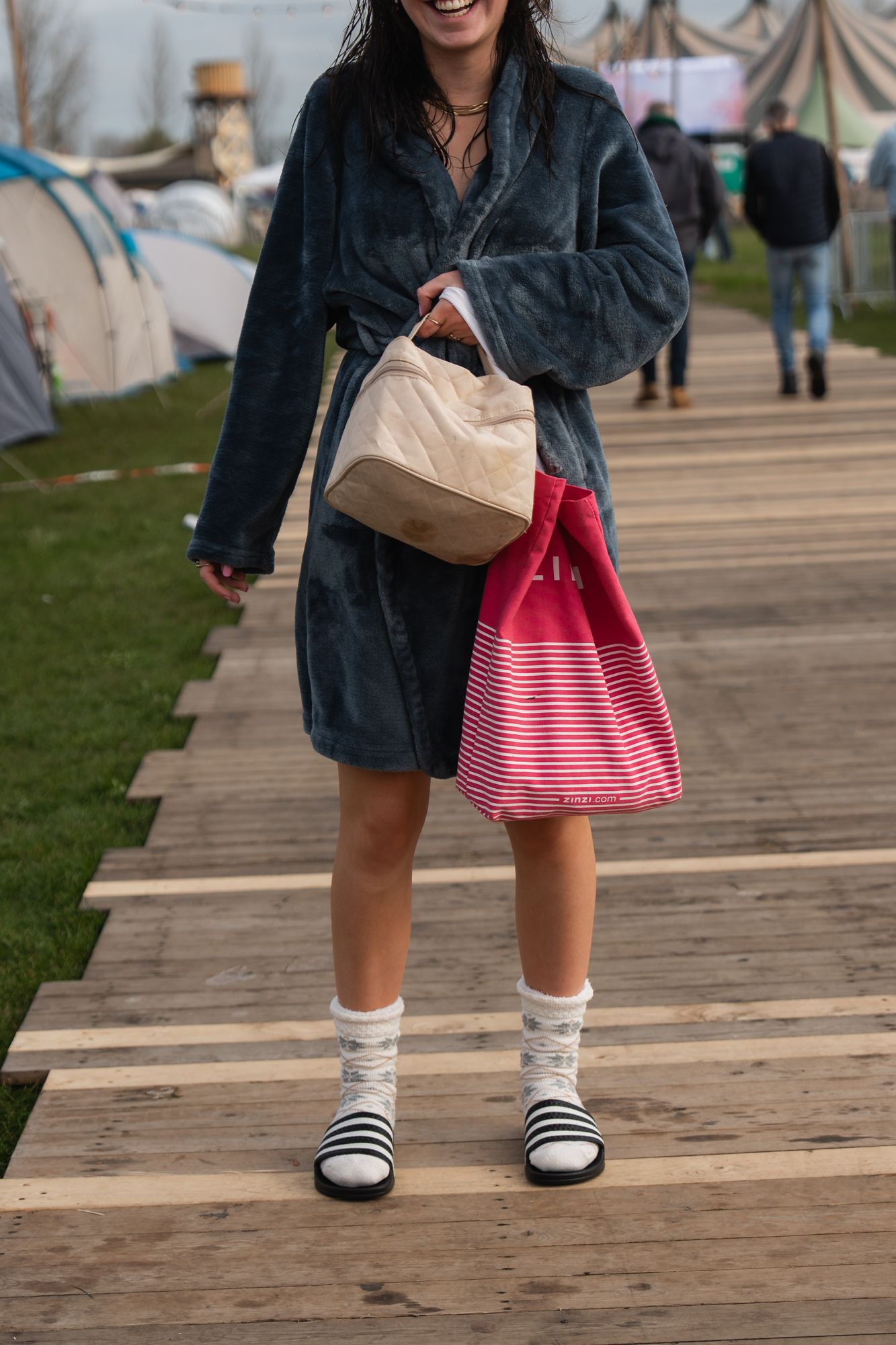 Woman in blue robe holding beige and pink striped bags, wearing white socks with black sandals on wooden walkway.