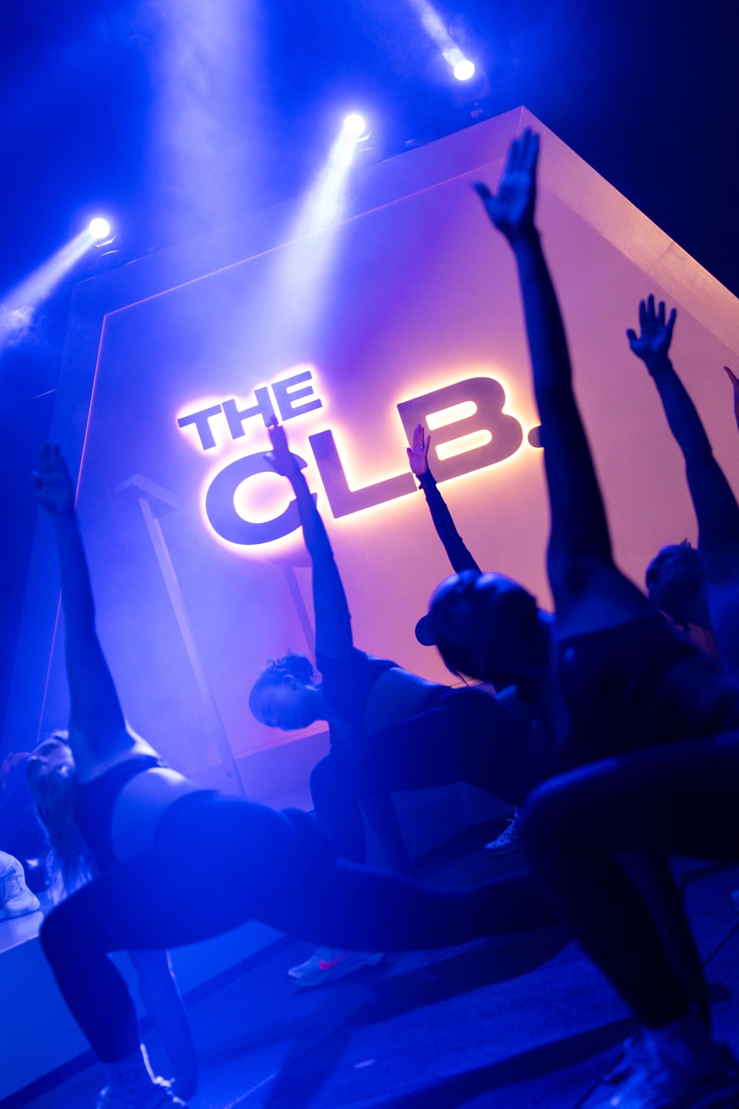 Silhouettes of people raising hands at The CLB fitness studio event, bathed in dramatic blue stage lighting.