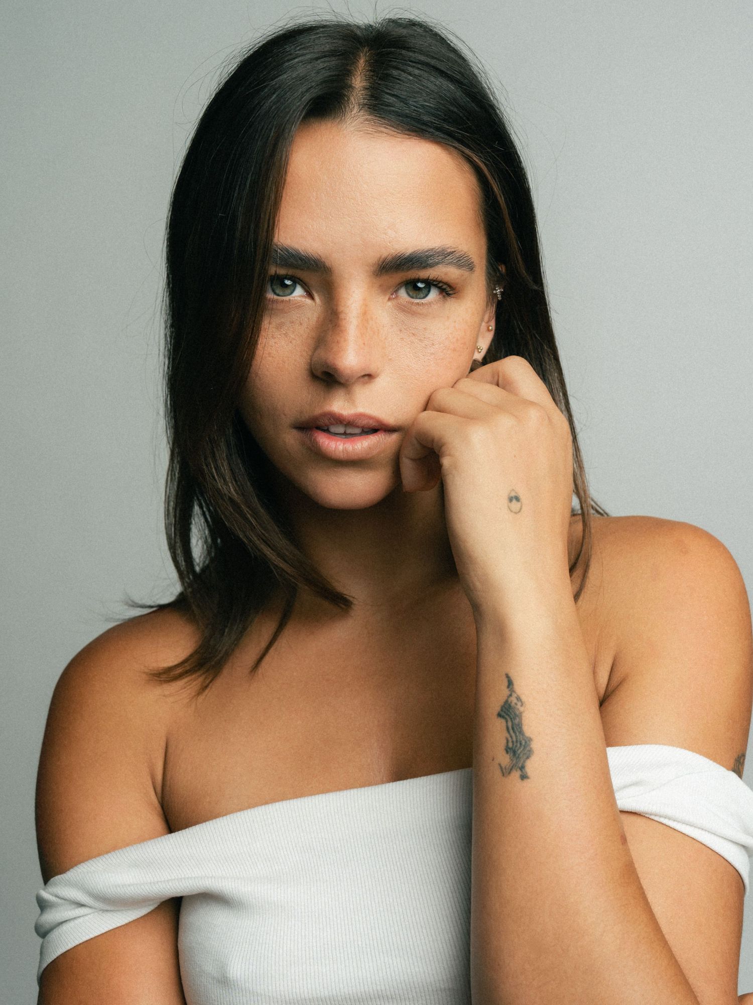 Young woman with dark hair, freckles, and a wrist tattoo poses in a white off-shoulder top against a gray background.