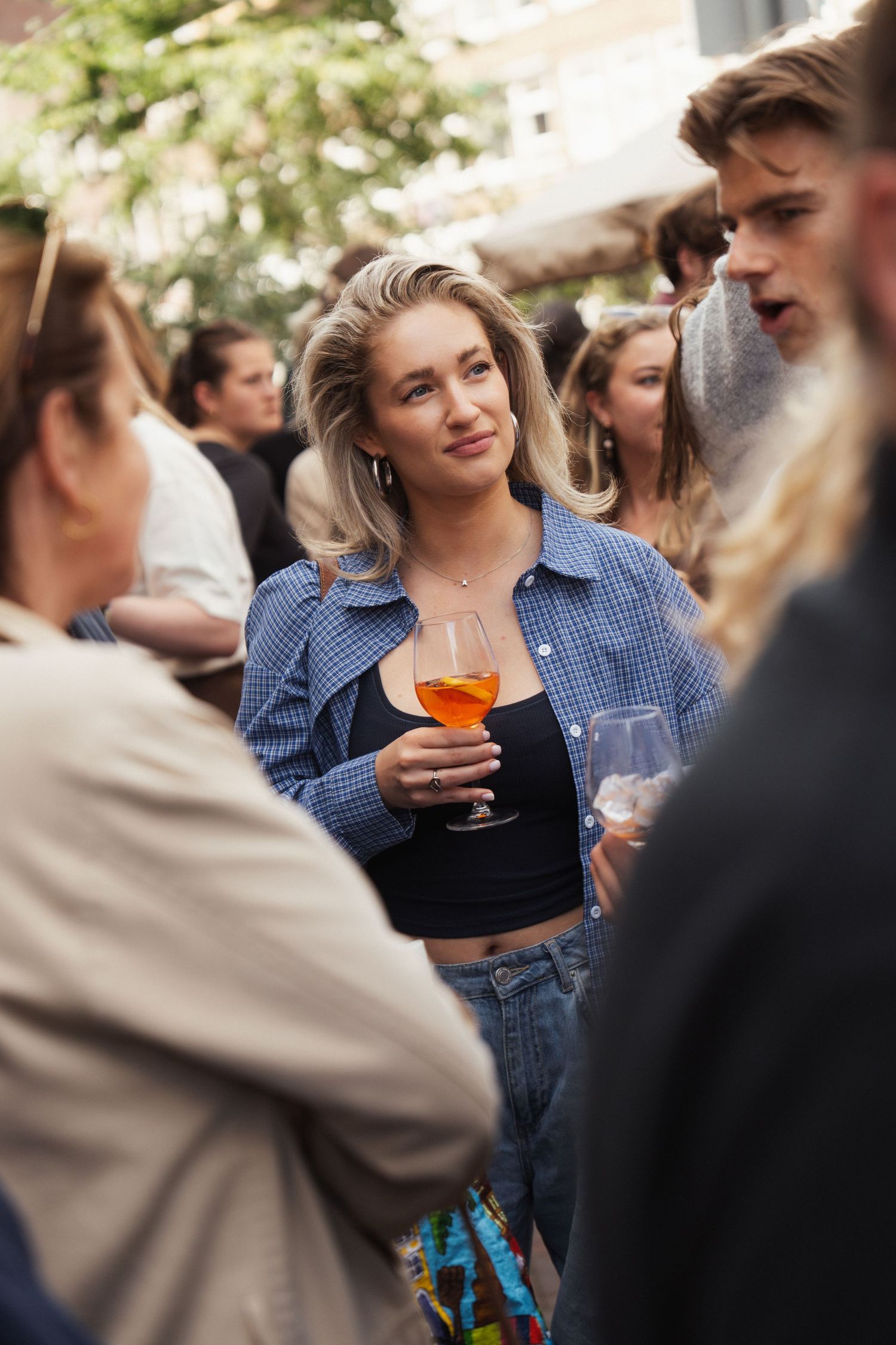 Blonde woman holding an orange cocktail at an outdoor social gathering, wearing a blue shirt and black crop top.