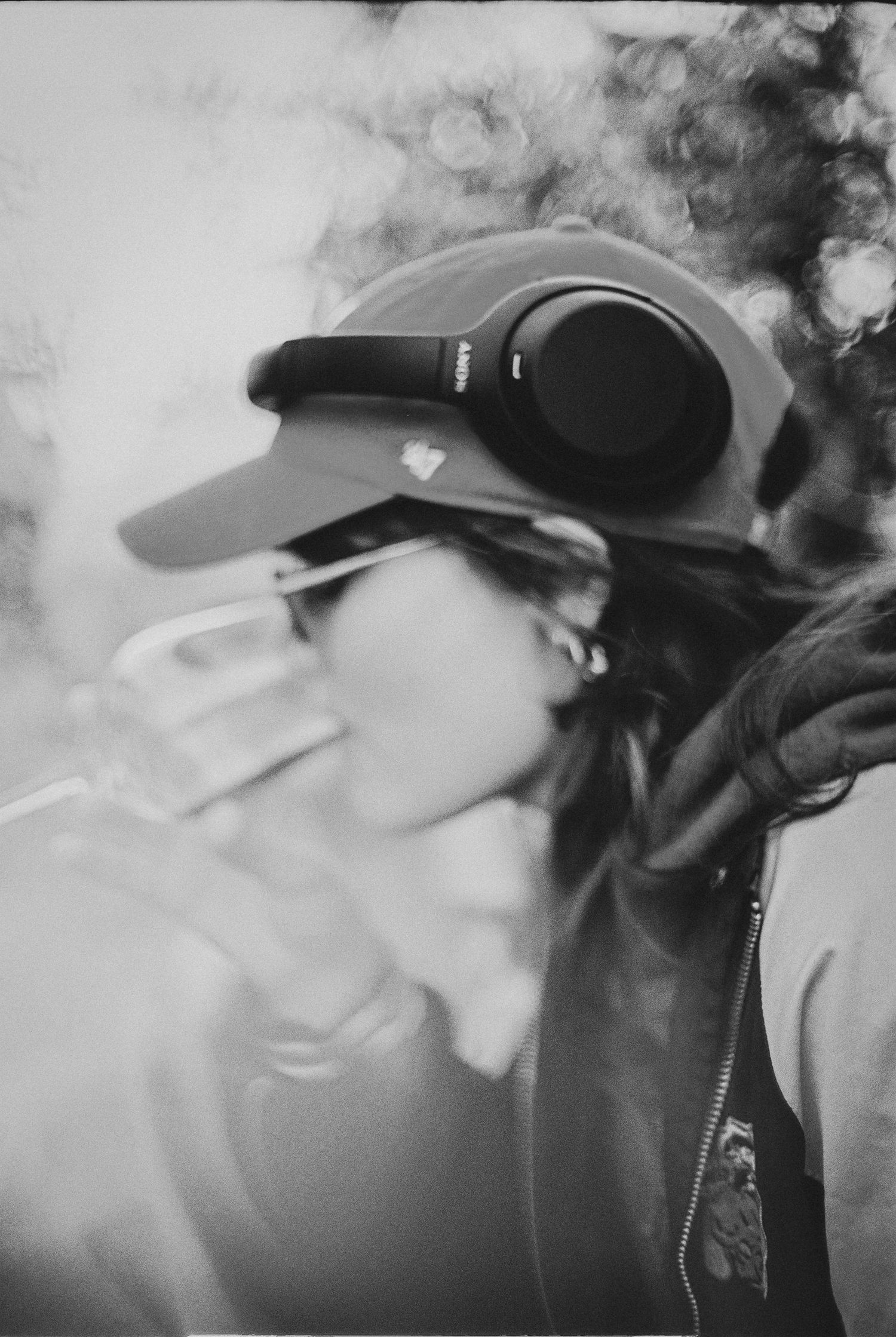 Black and white photo of a woman wearing headphones and a cap, drinking from a cup outdoors.