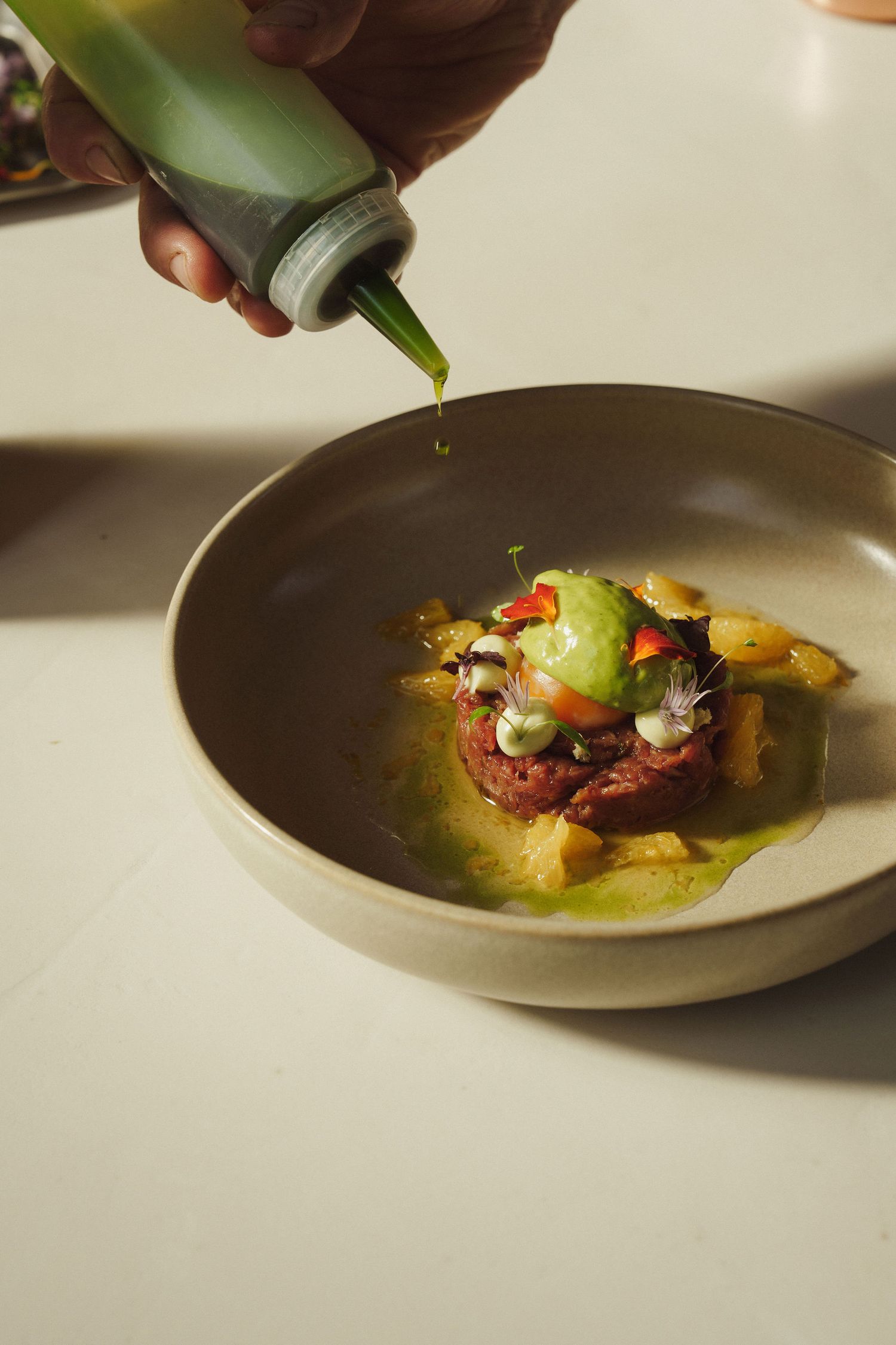 Chef drizzling green oil over a gourmet beef tartare with avocado and edible flowers in a ceramic bowl.