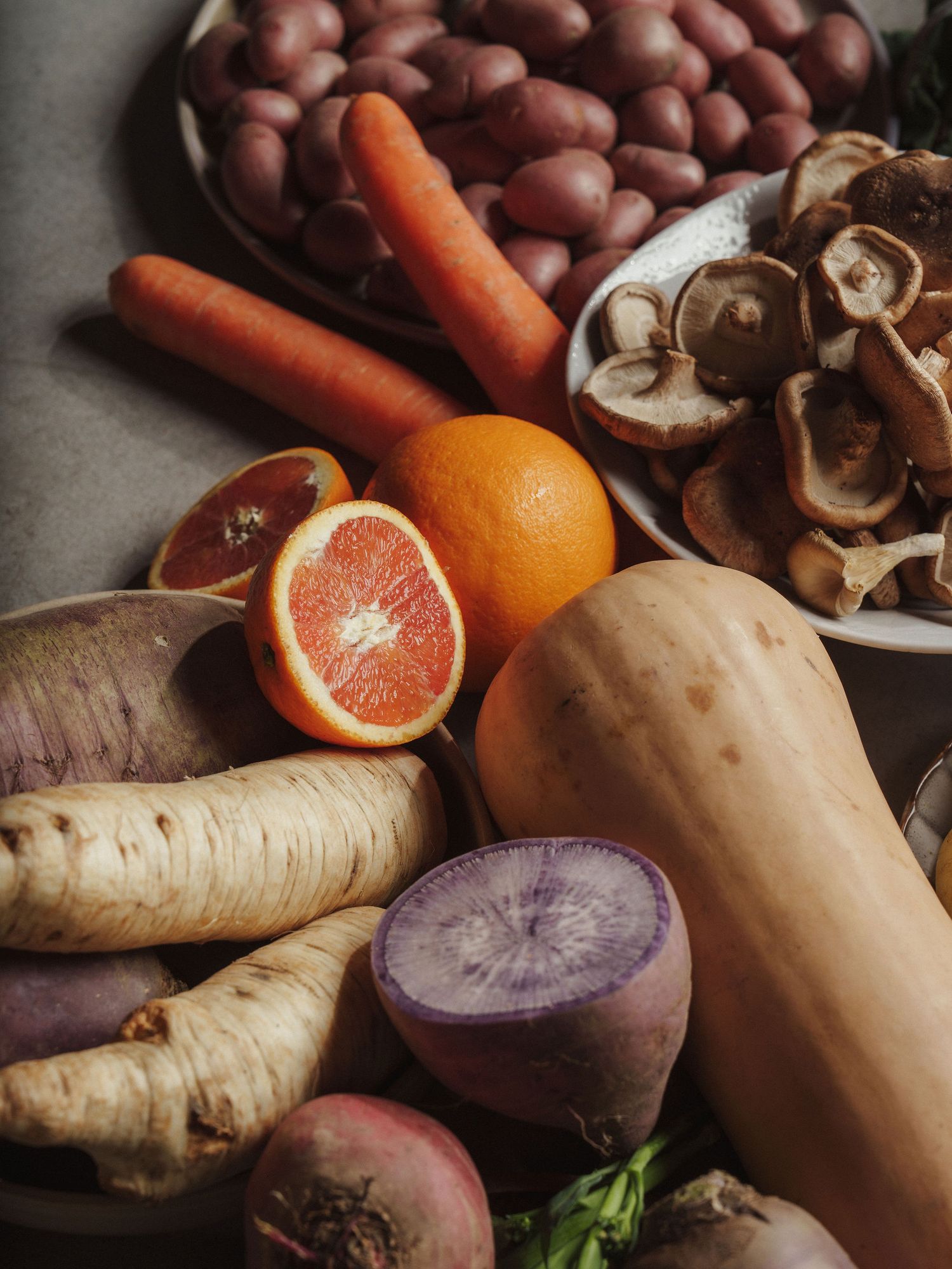 Assorted fresh vegetables and fruits including carrots, butternut squash, beets, parsnips, mushrooms, potatoes, and sliced grapefruit.