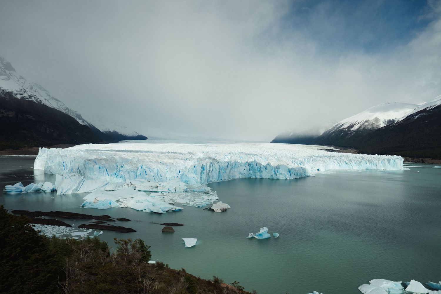 Massive Perito Moreno glacier meeting turquoise waters with floating icebergs, surrounded by snow-capped Patagonian mountains.
