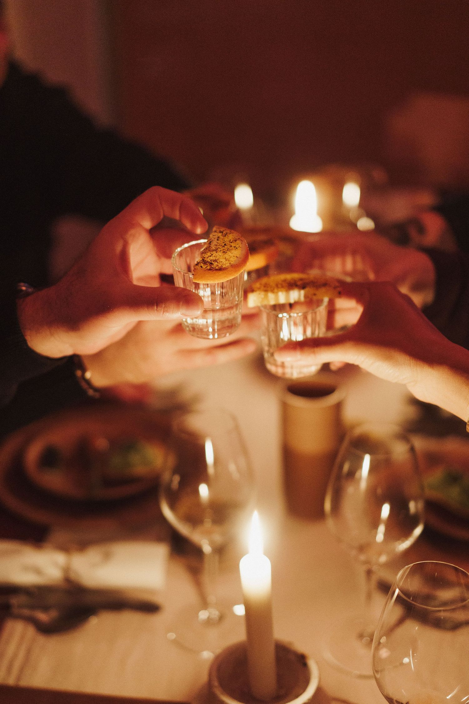 Hands clinking shot glasses over a candlelit dinner table with wine glasses and warm ambient lighting.