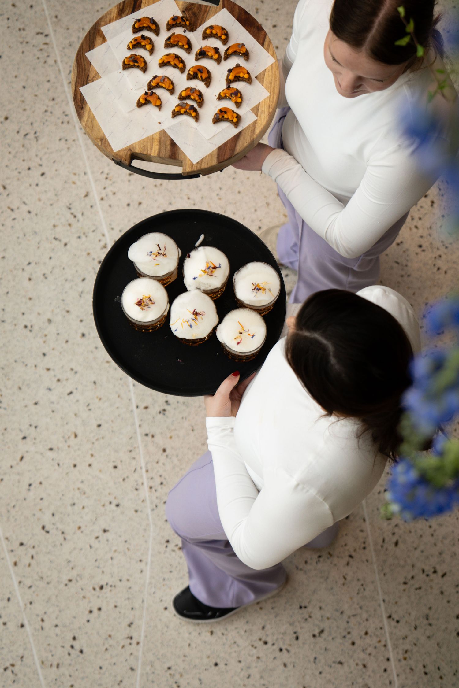 Two servers in white tops hold trays of elegant desserts and canapes at a catering event.