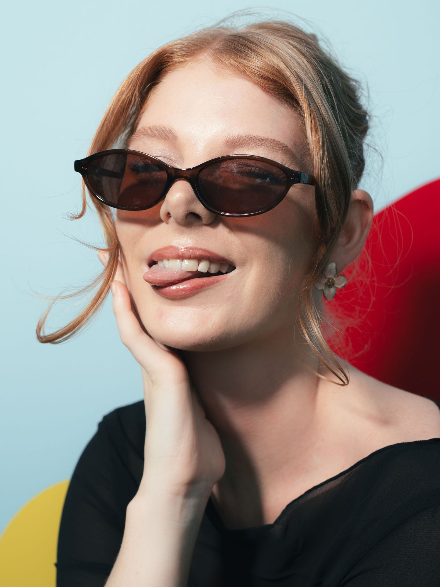 Smiling redhead woman wearing brown cat-eye sunglasses and a black top, resting hand on cheek against a blue background.