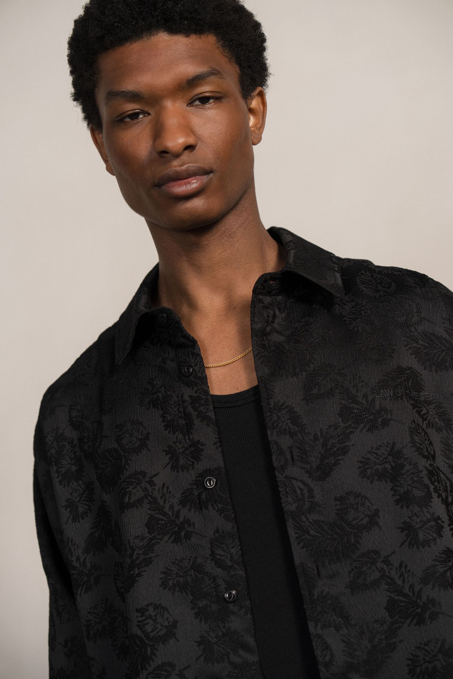 A fashion portrait showing a person wearing a black floral jacquard button-up shirt over a dark t-shirt against a neutral background.