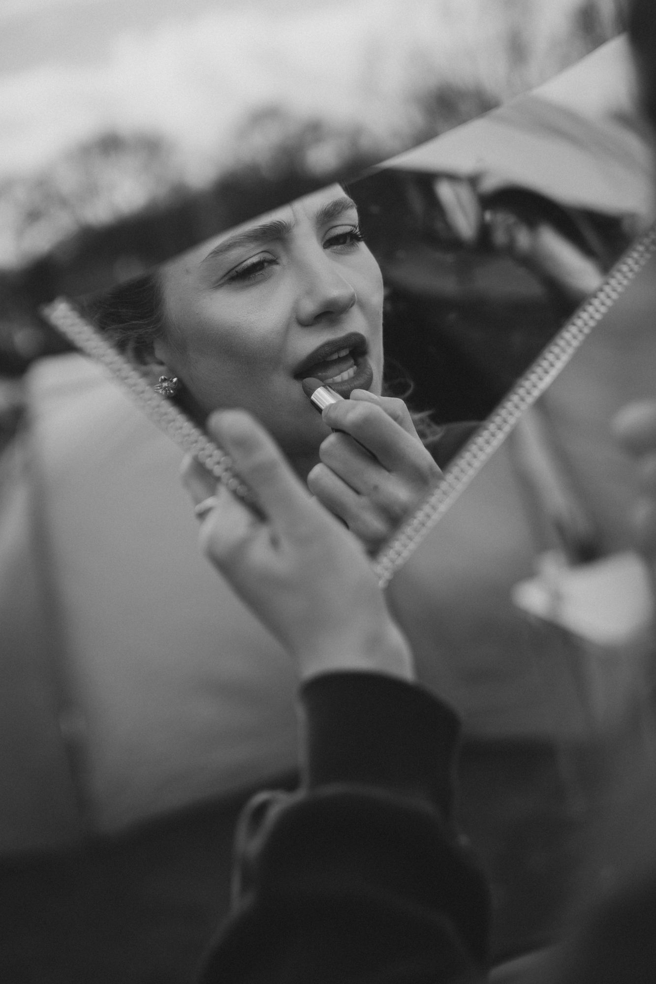Black and white photo of a woman applying lipstick, reflected in a broken mirror outdoors.