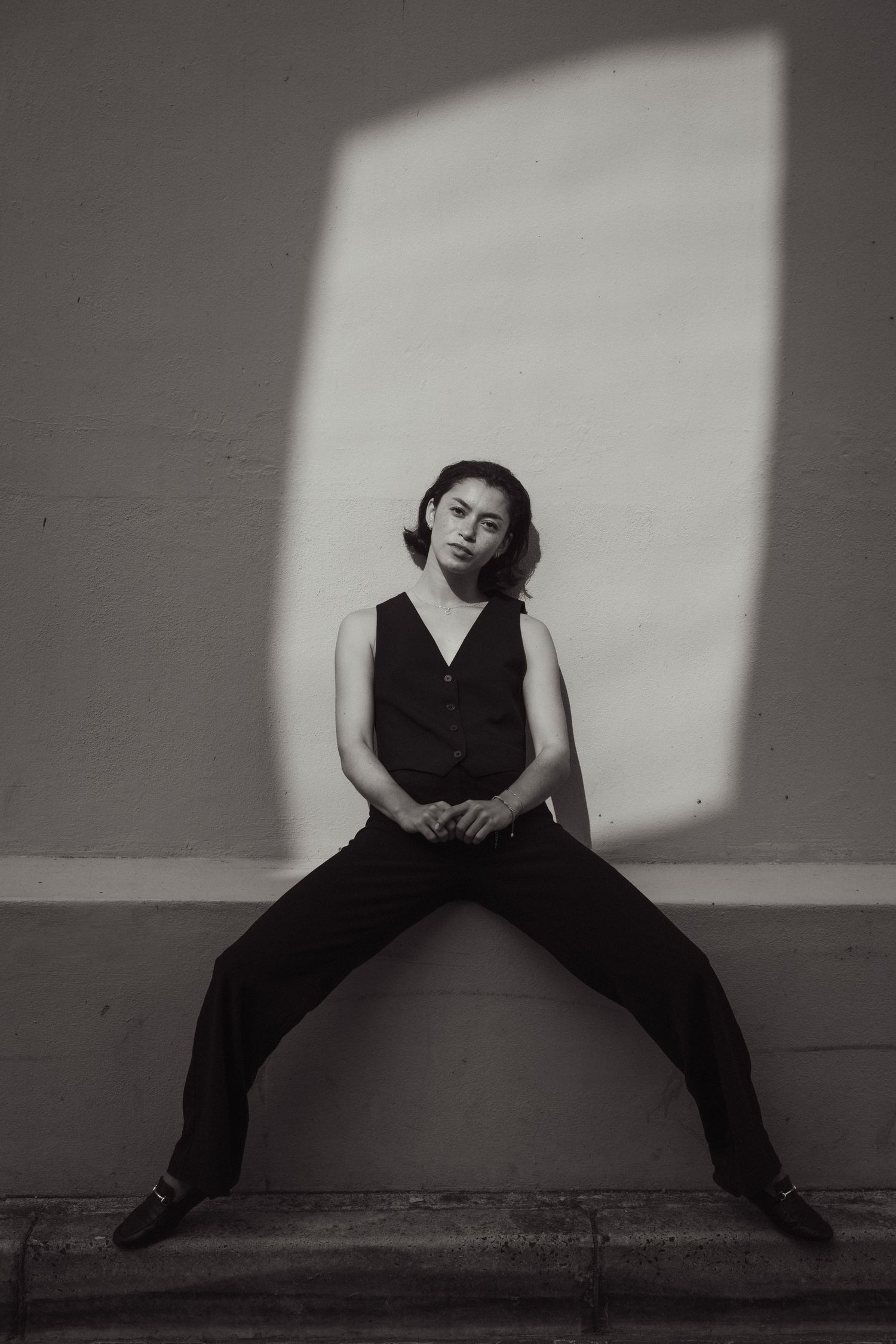 Black and white portrait of a young woman in a vest and trousers seated against a wall with dramatic light.