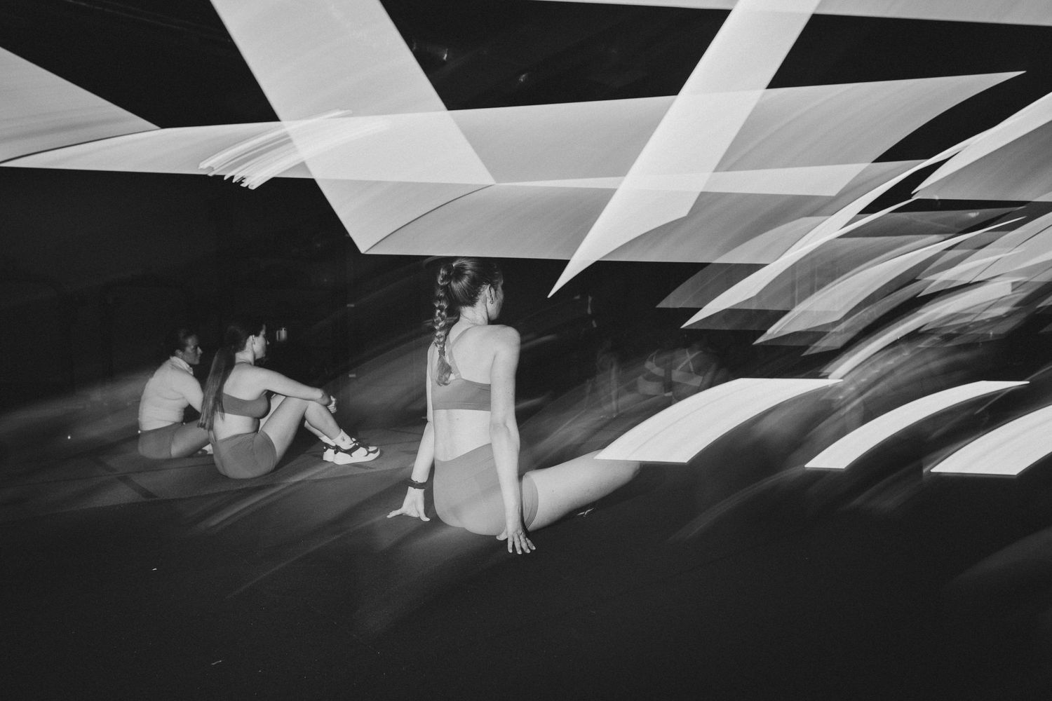 Two women resting on floor under geometric light projections in black and white fitness setting.