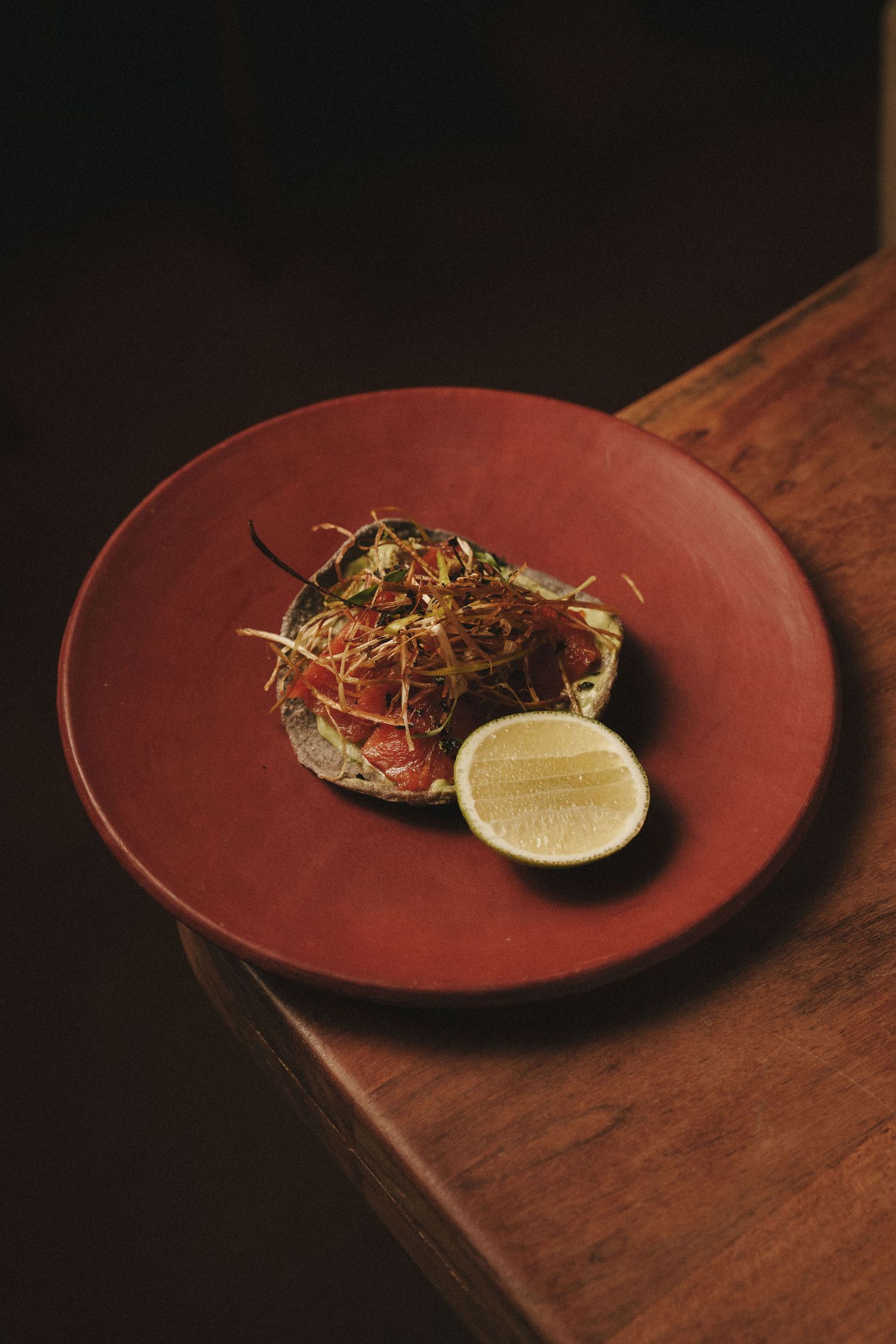 Elegant plated dish with fresh fish, crispy garnish, and lime half on a red ceramic plate atop a wooden table.