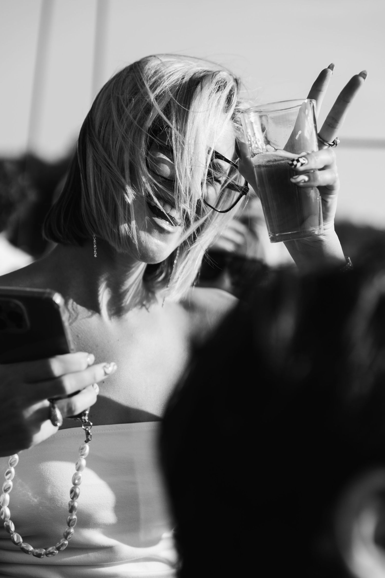 Black and white photo of a blonde woman in glasses sipping a drink while holding a smartphone outdoors.