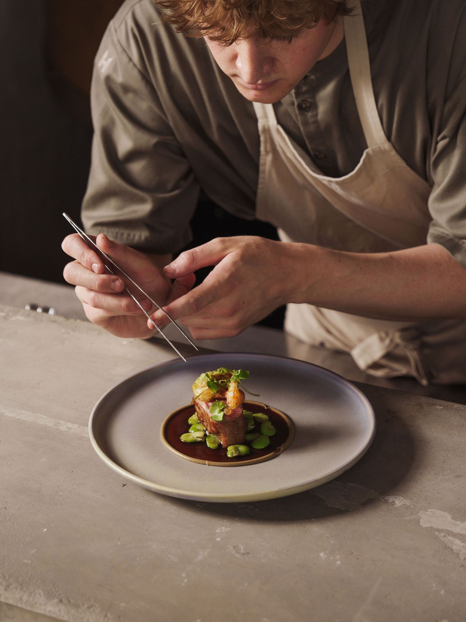 Chef carefully plates an elegant fine dining dish with tweezers on a white ceramic plate in a restaurant kitchen.