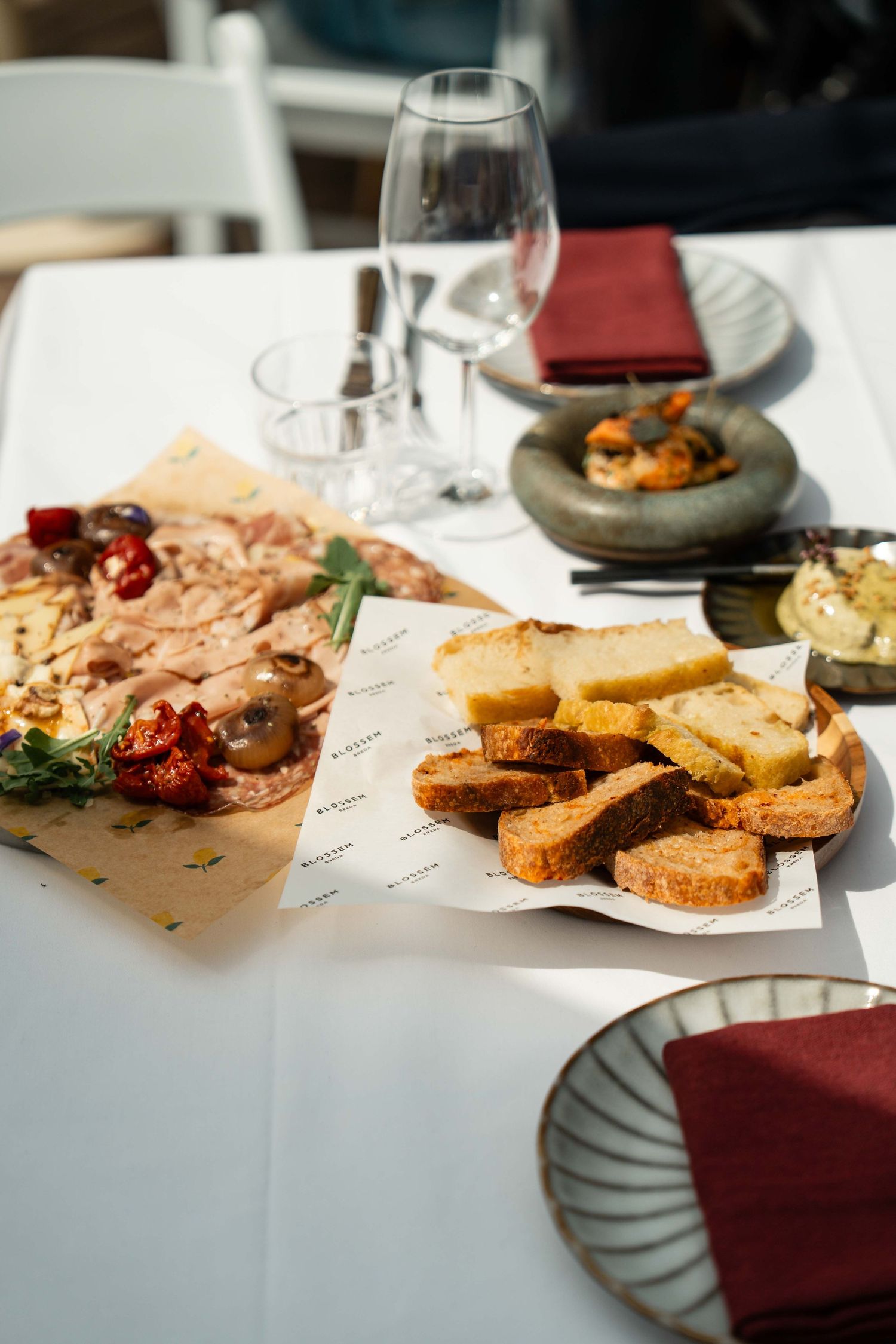 Elegant restaurant table with charcuterie board, toasted bread slices, wine glasses, and red napkins in bright sunlight.