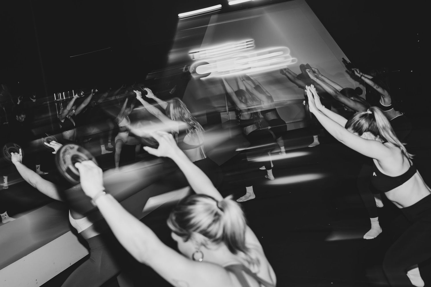 Black and white photo of women exercising in a dark fitness studio with neon lights in the background.
