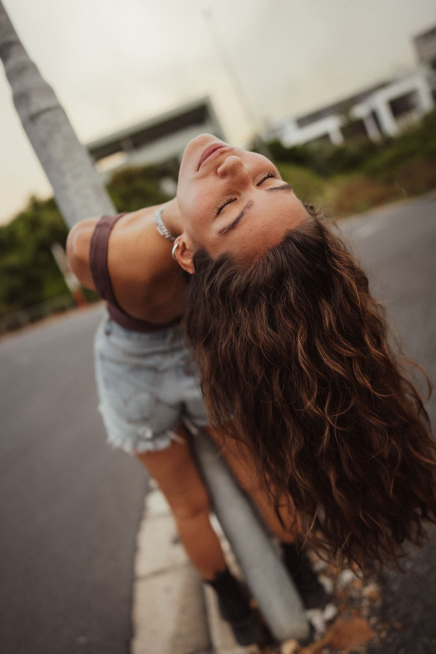 Young woman with curly hair leaning back against a pole outdoors, wearing denim shorts and a brown top.