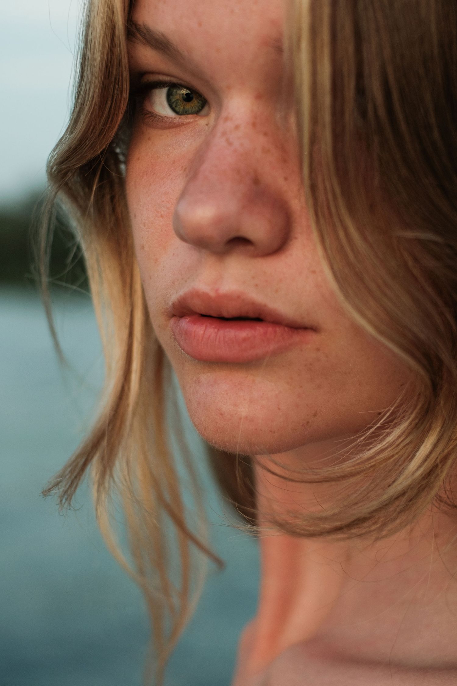 Close-up portrait of a young woman with freckles, green eyes, and blonde hair outdoors near water.