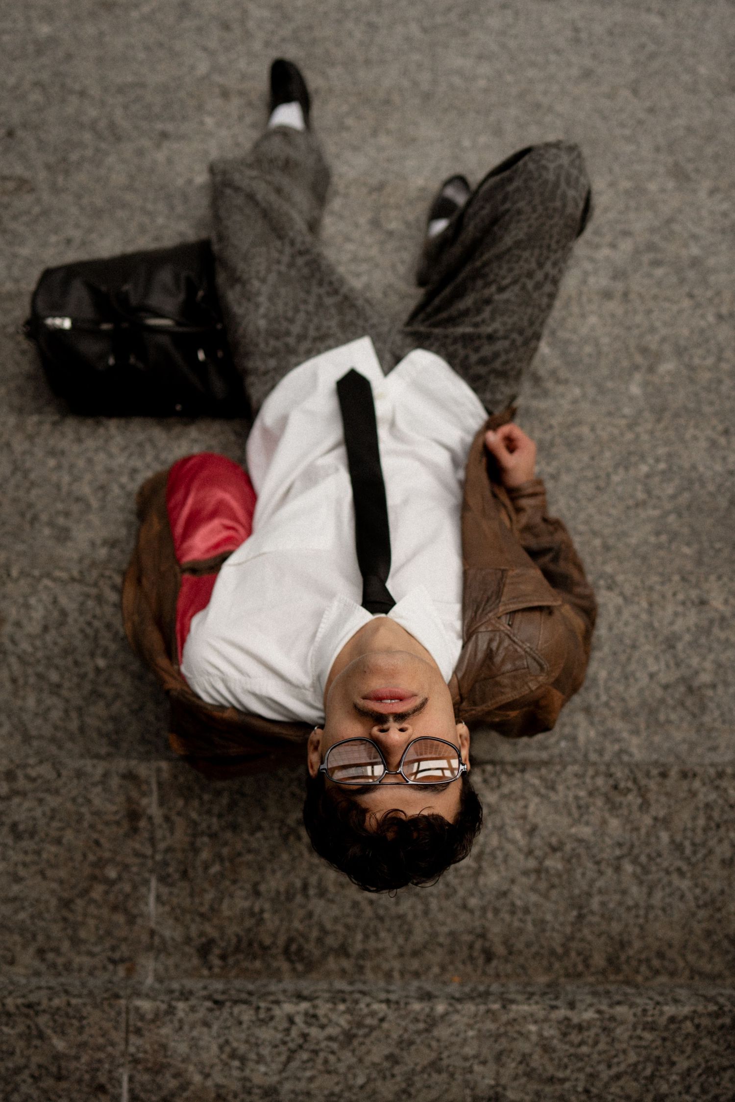 Businessman lying on floor with glasses, briefcase nearby, wearing white shirt and tie.