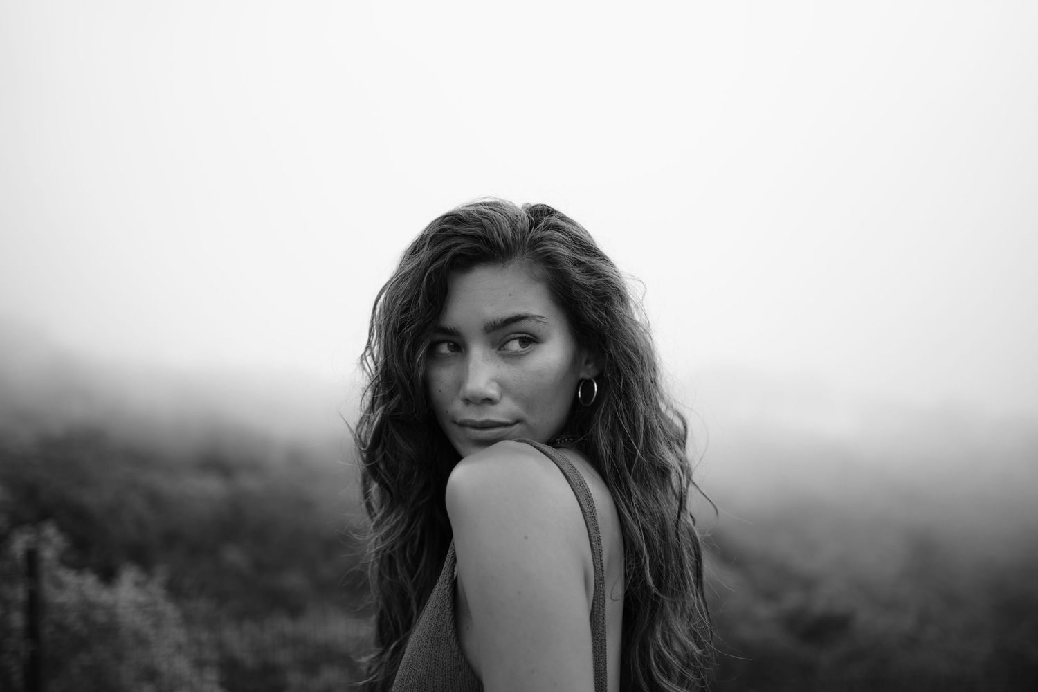 Black and white portrait of a young woman with wavy hair gazing over her shoulder in a misty outdoor setting.