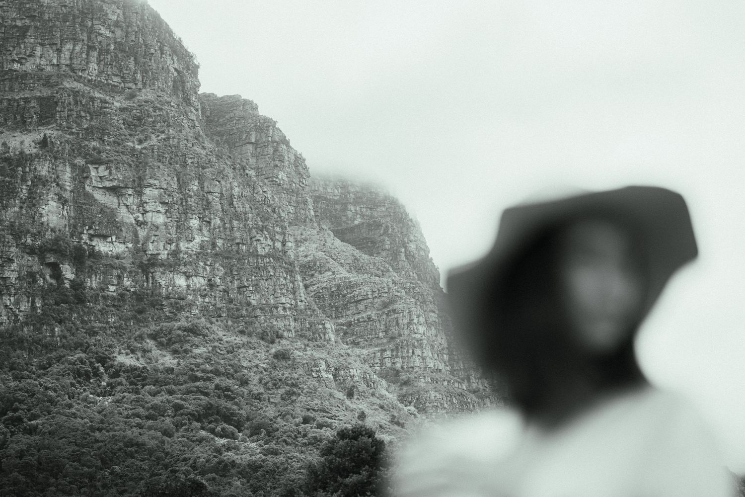 Black and white photo of a blurred figure in a hood with a dramatic rocky mountain and lush greenery in the background.