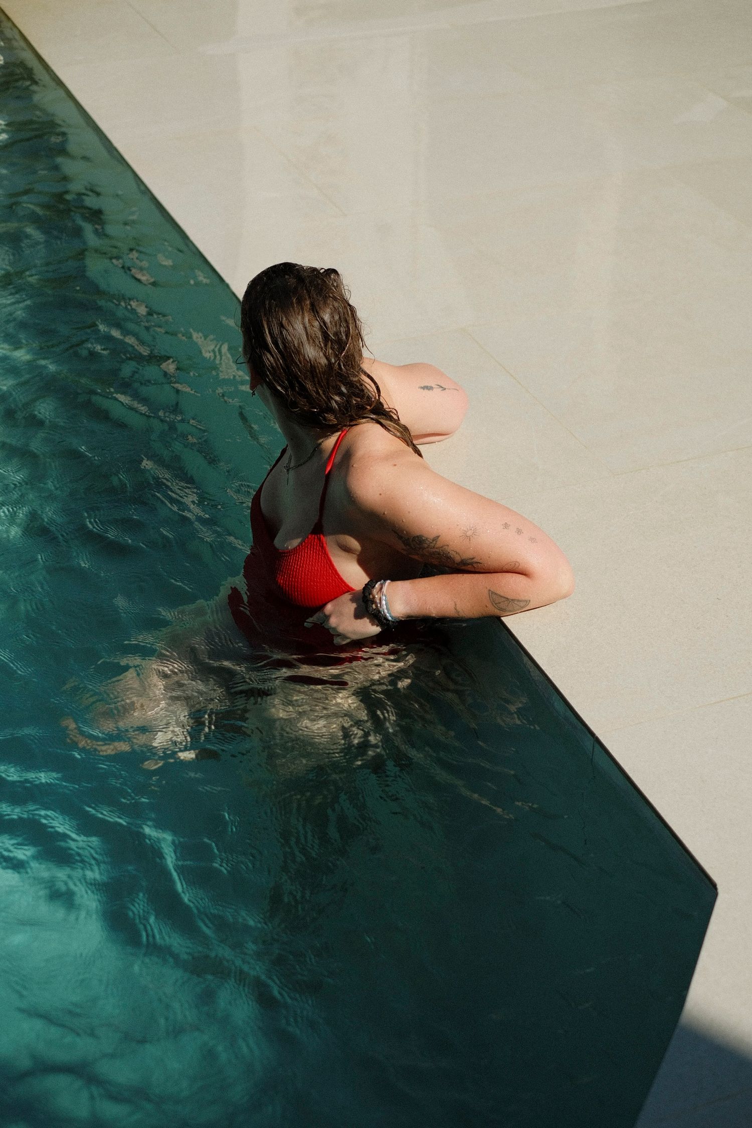 Woman in red bikini resting at the edge of a turquoise swimming pool, viewed from above.