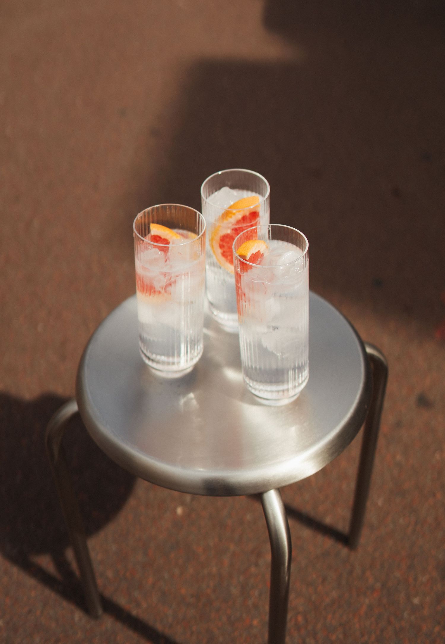 Three iced drinks with grapefruit slices in tall glasses on a silver metal stool table outdoors in sunlight.