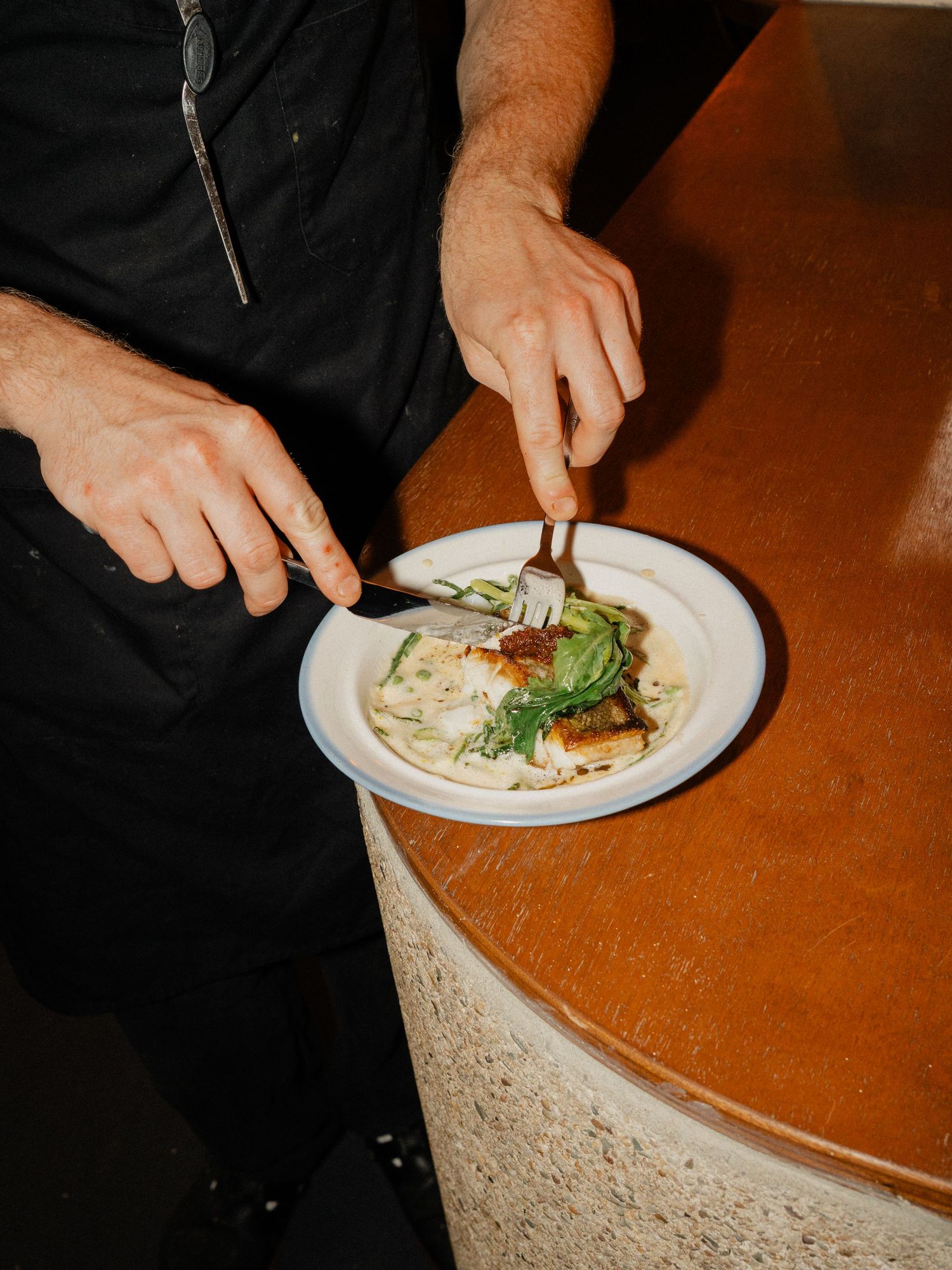 Chef in black uniform plating a gourmet dish with greens on a white plate at a bar.