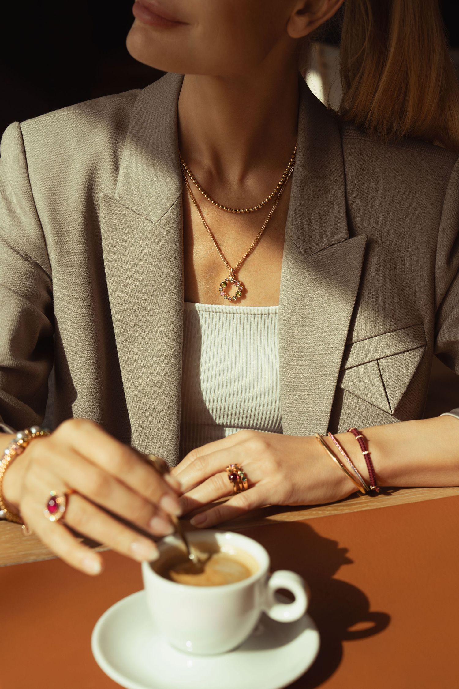 Woman in beige blazer wearing layered gold necklaces, ruby rings and bracelet, stirring espresso coffee cup.