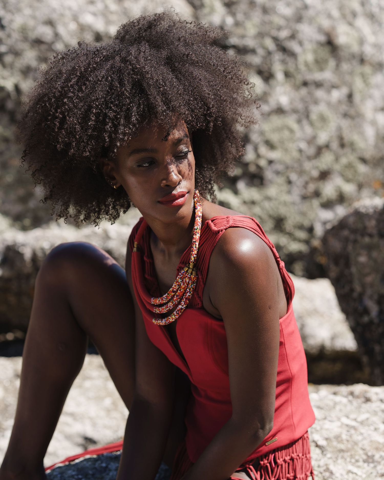 Young Black woman with natural afro hair wearing red top and beaded necklace, sitting on rocks outdoors.