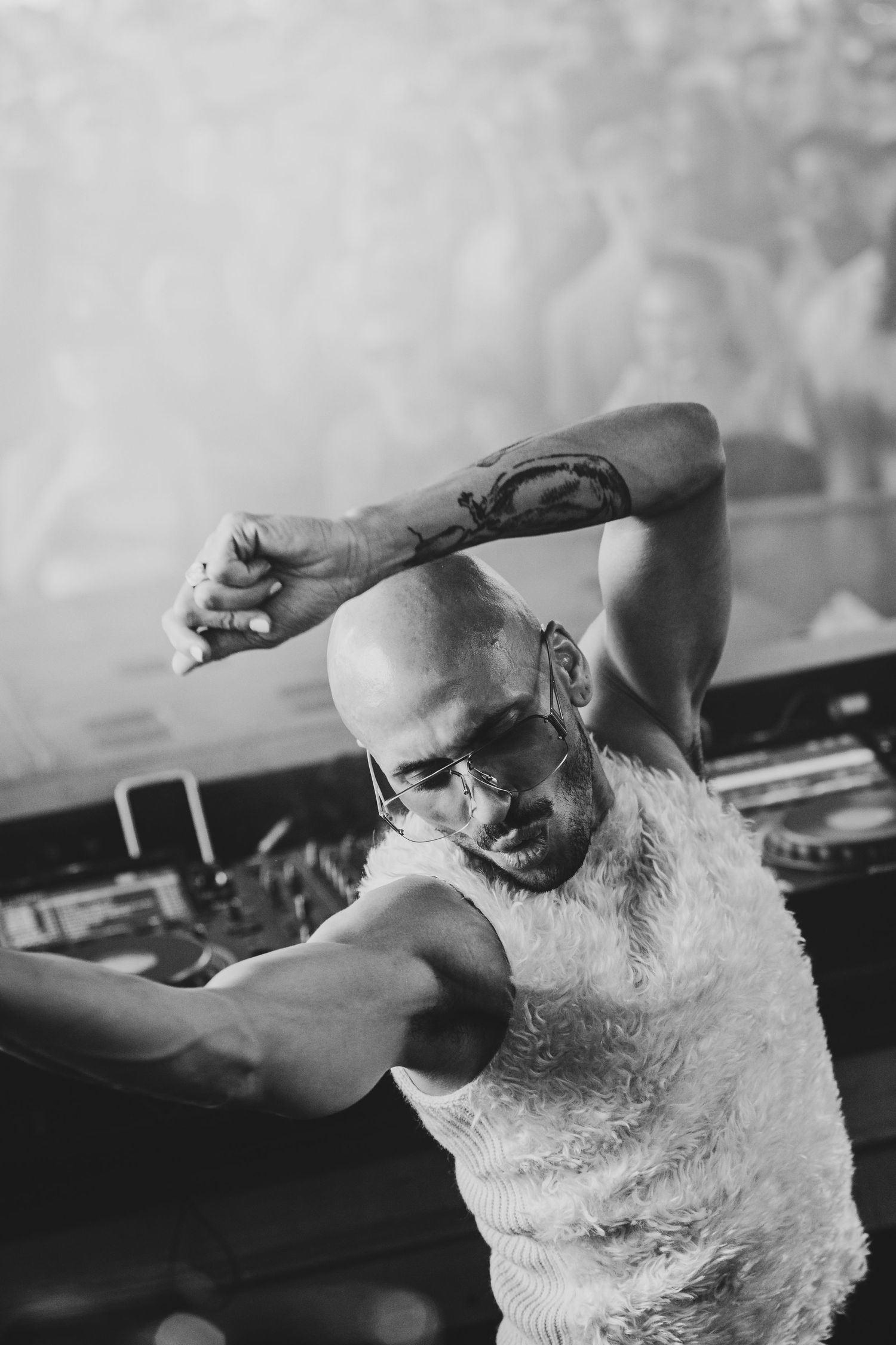 Bald DJ with tattoos and glasses dancing energetically at turntables in black and white photo.