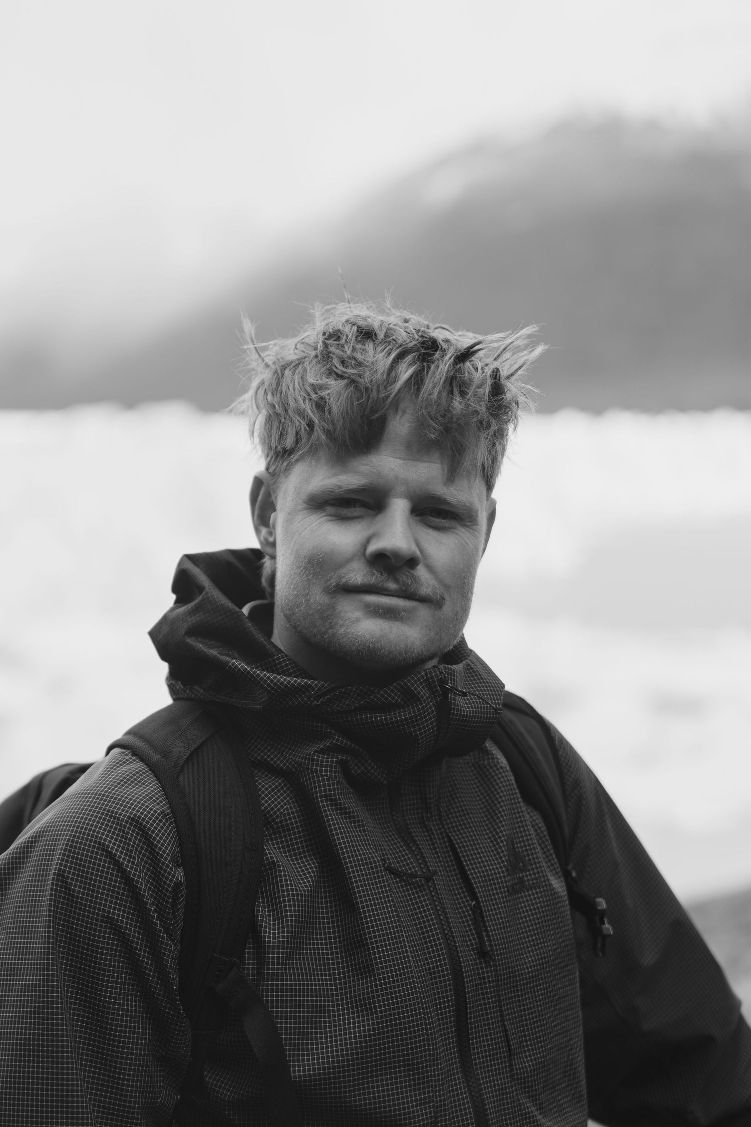 Black and white portrait of a young man with tousled hair wearing a rain jacket outdoors with misty mountains behind.