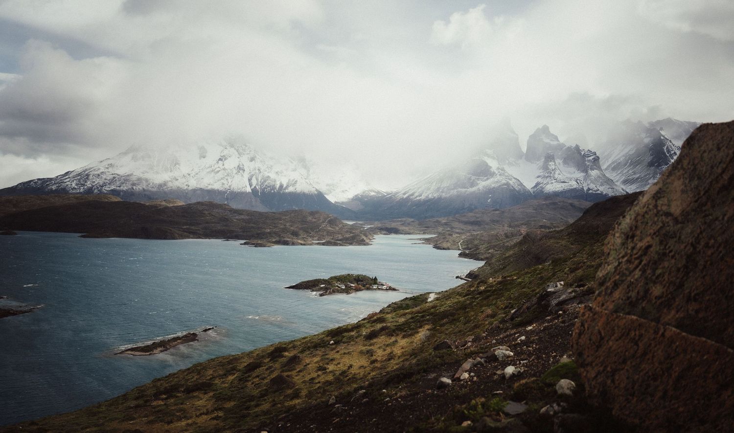 Snow-capped mountains overlook a serene blue lake surrounded by rugged terrain under cloudy skies in Patagonia.