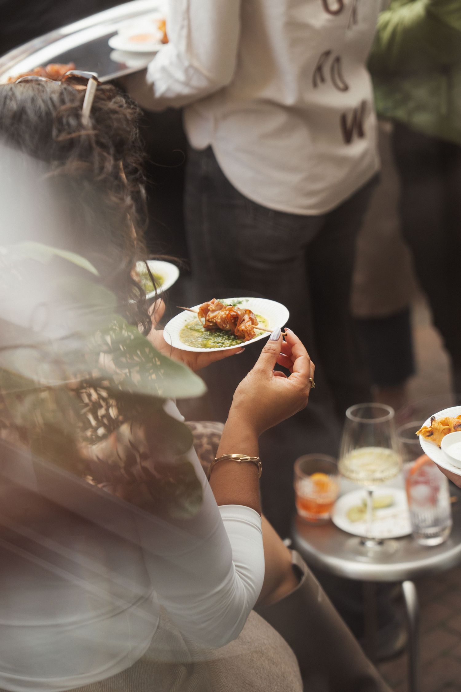 Woman holding a small plate of food at an outdoor gathering with drinks and appetizers on a metal table.