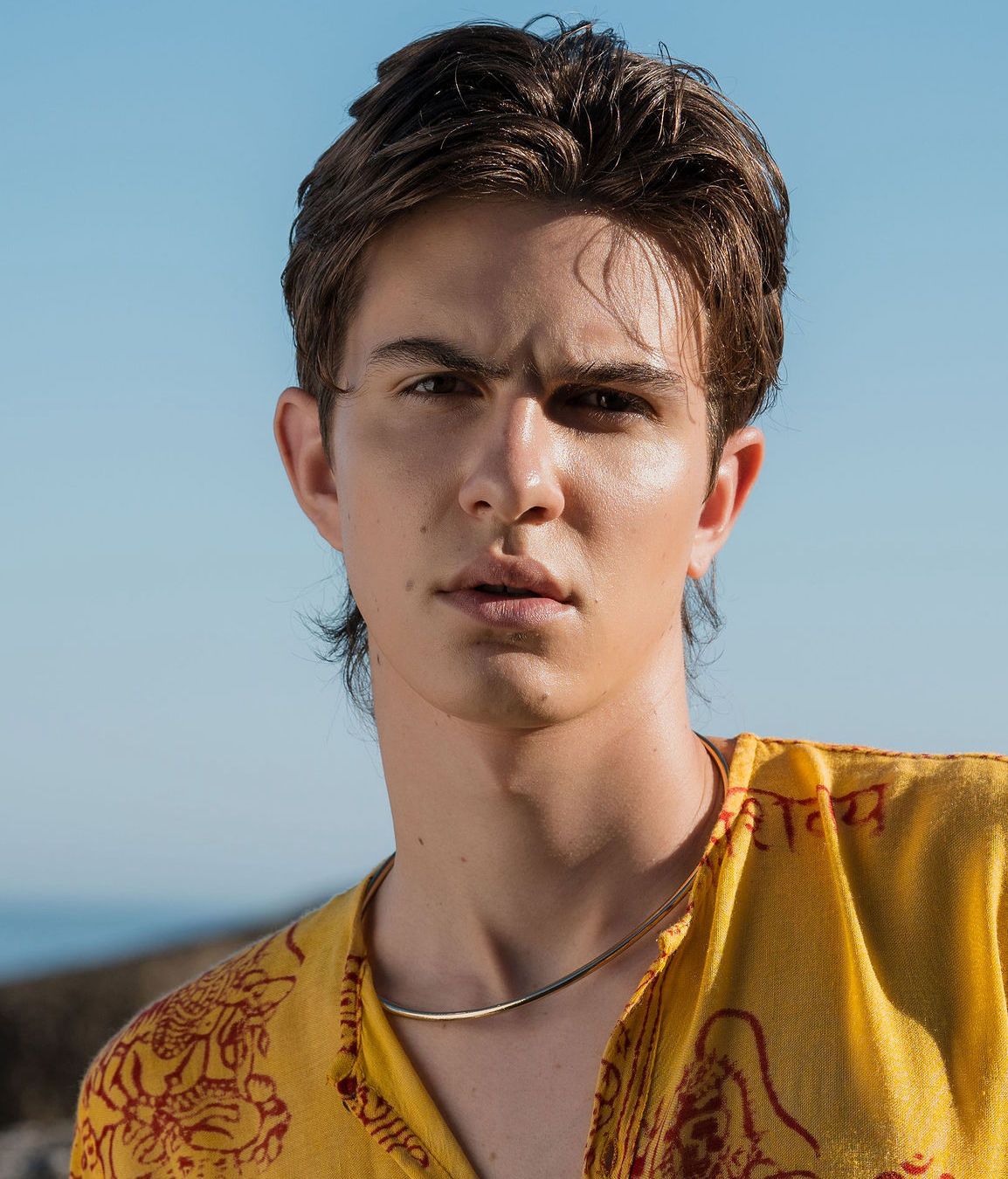 Young man with dark hair wearing a yellow patterned shirt and silver necklace, posing outdoors against a blue sky.