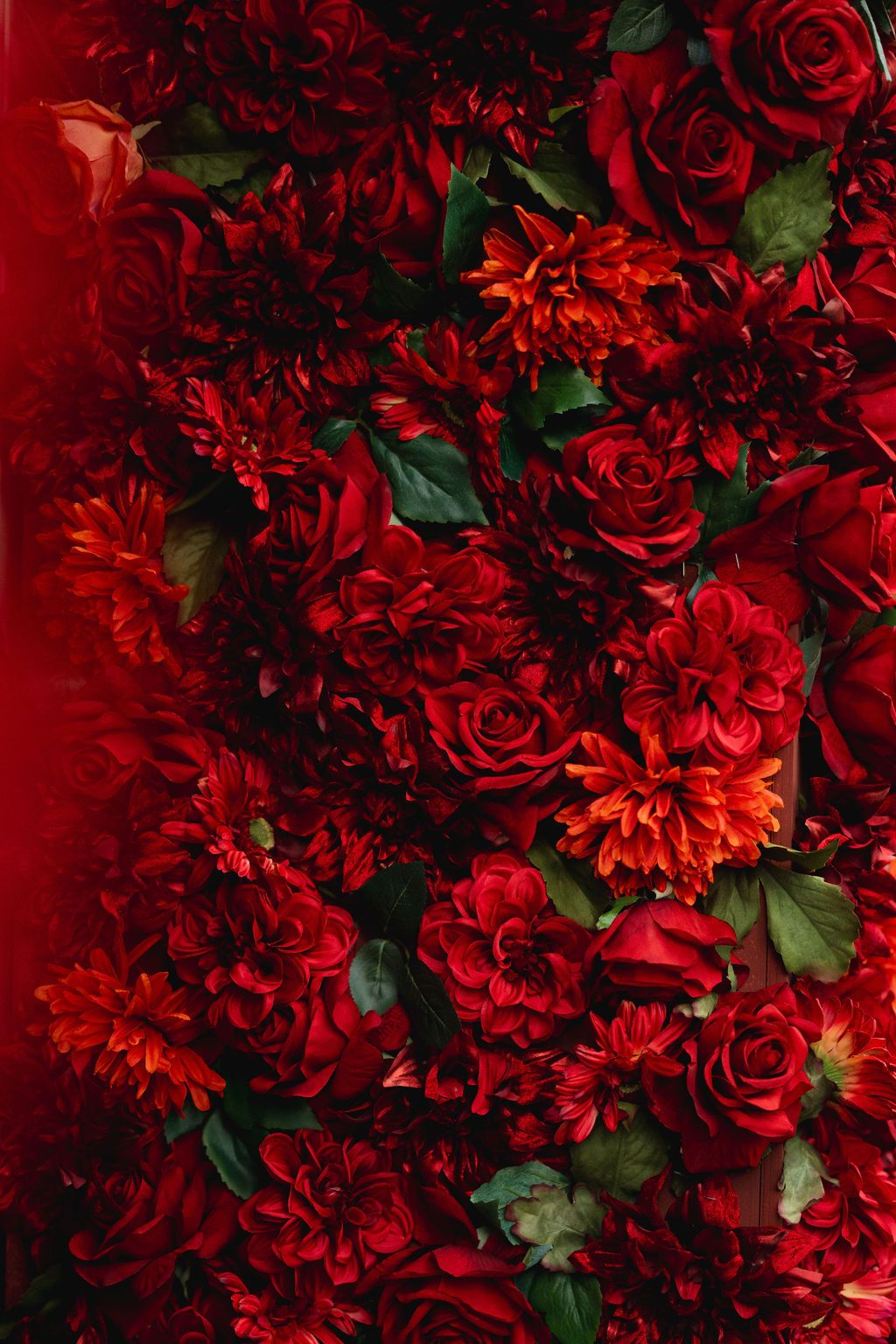 Lush arrangement of deep red roses, dahlias, and carnations with green leaves creating a rich floral wall display.