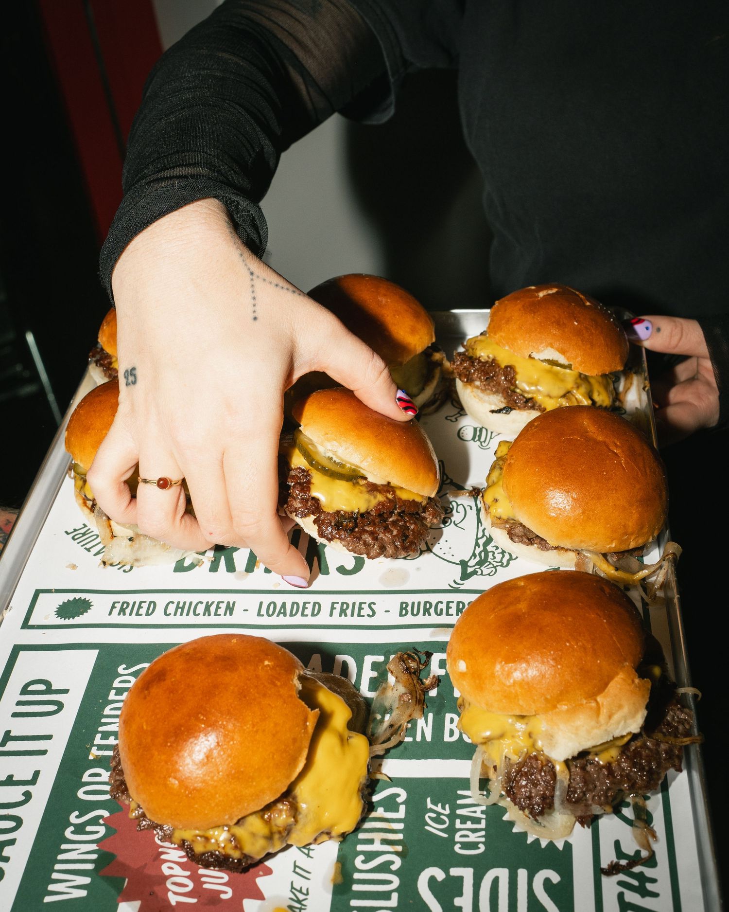 A hand with red nails reaches for mini cheeseburgers on a tray at a burger restaurant.