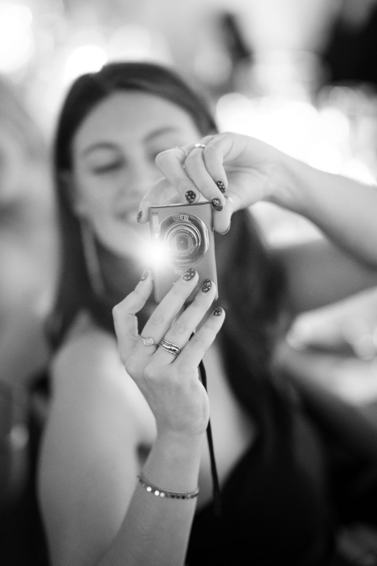 Black and white photo of a young woman taking a selfie with a compact camera, wearing rings and a bracelet.