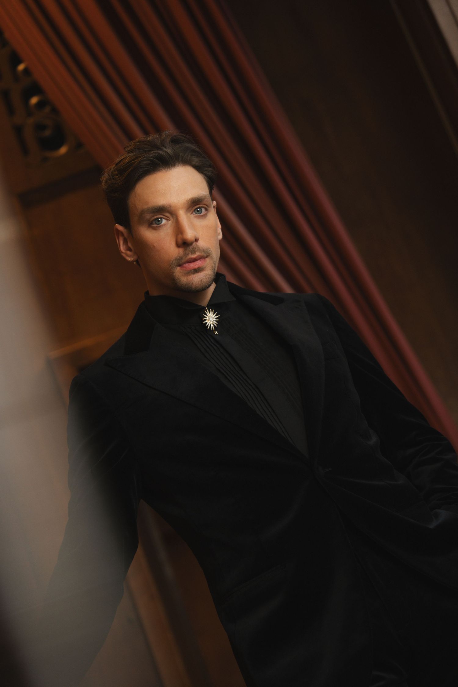 A stylish man in a black velvet suit with a star brooch poses elegantly against a warm, draped backdrop.