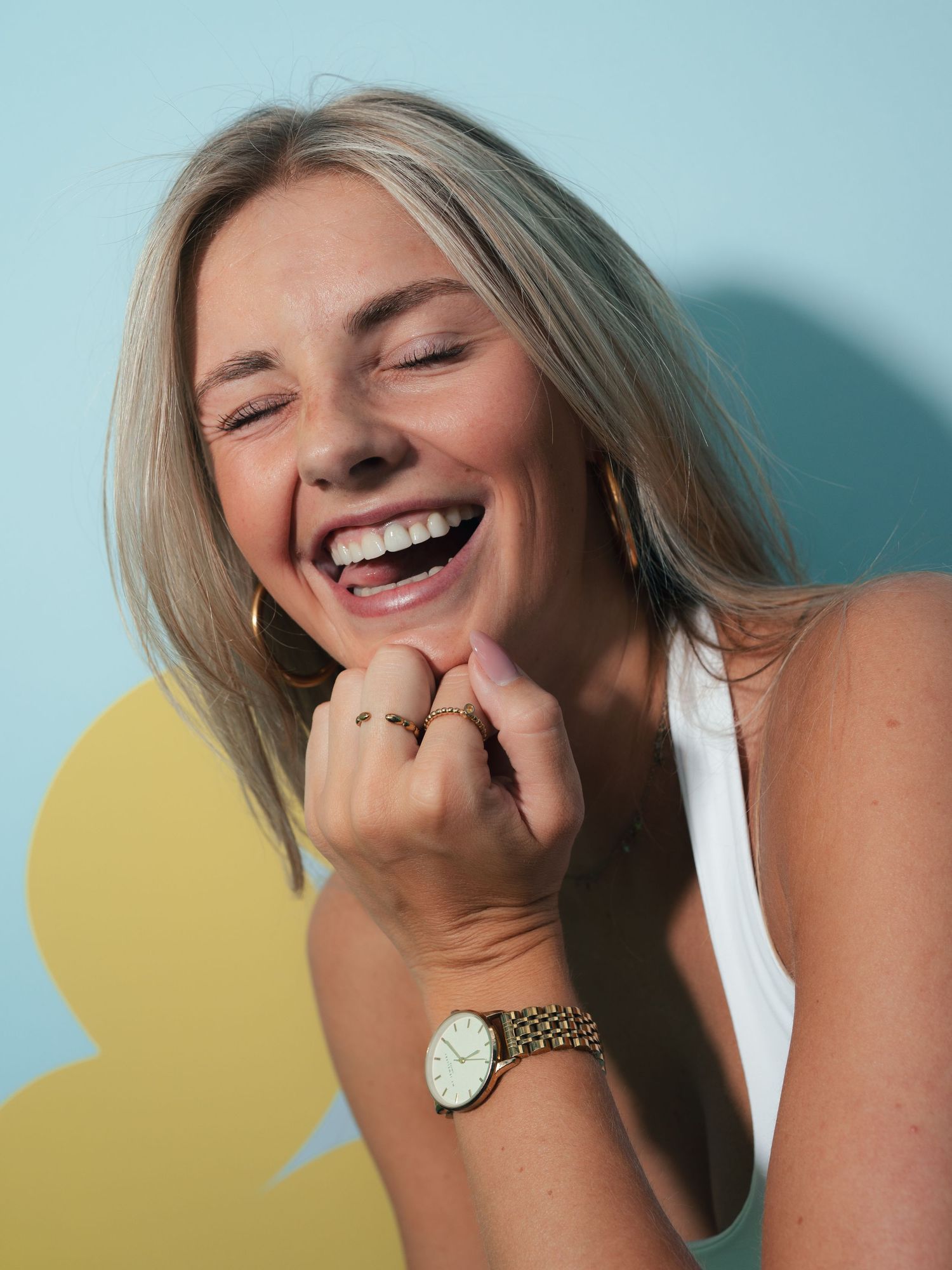 Blonde woman laughing joyfully wearing a white top and gold watch with hoop earrings against a teal background.
