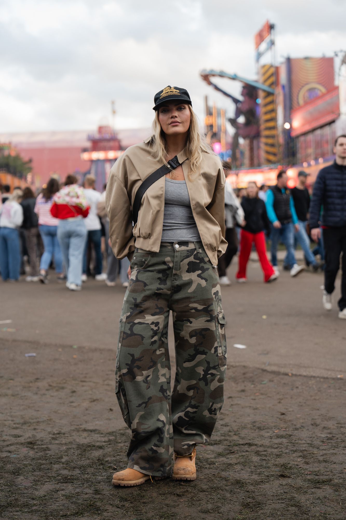 Woman in camo cargo pants, beige jacket, and cap poses at outdoor festival with crowd and rides behind her.